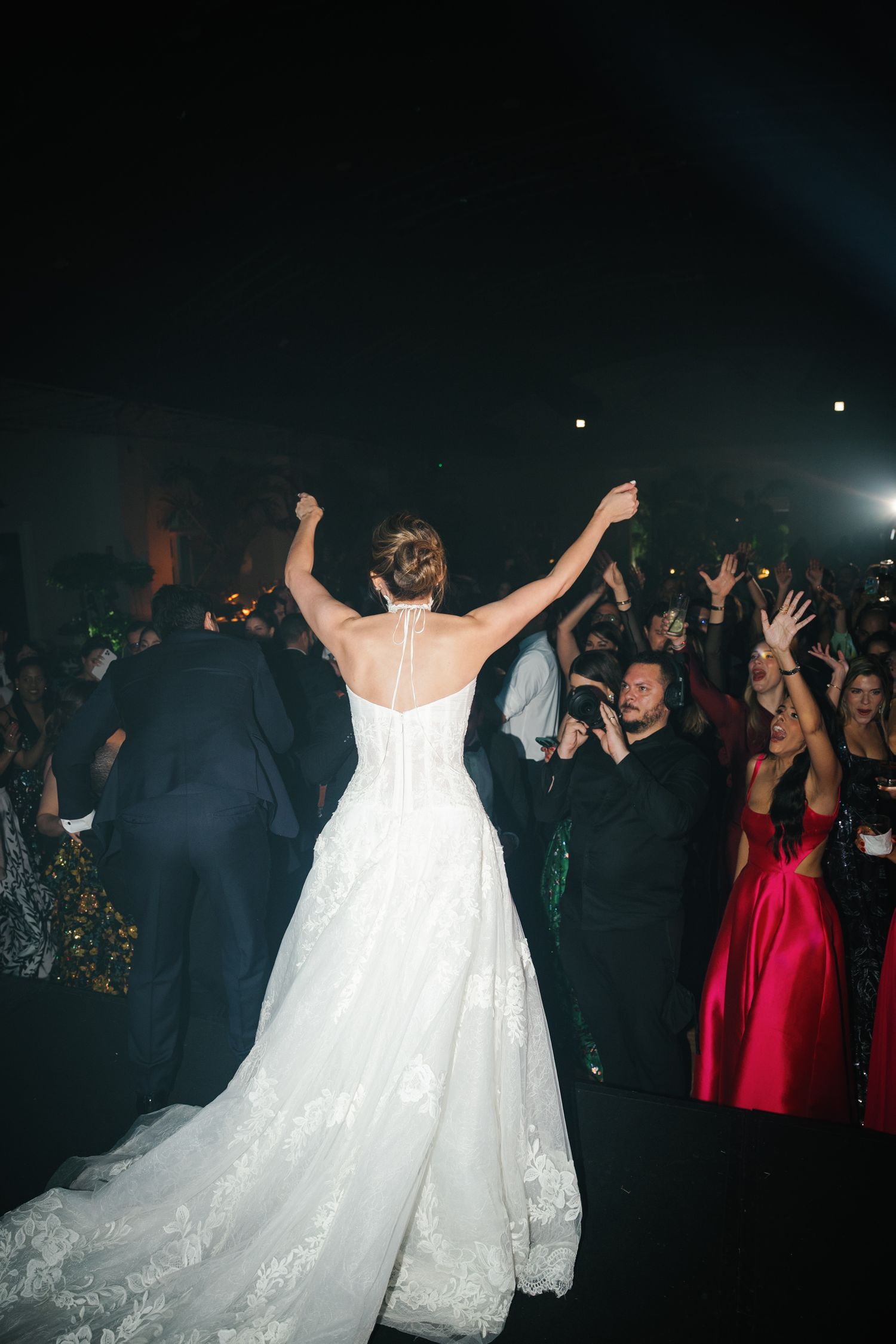 Wedding guests dance and celebrate on a dark dance floor while a bride in a flowing white gown raises her arms in joy.