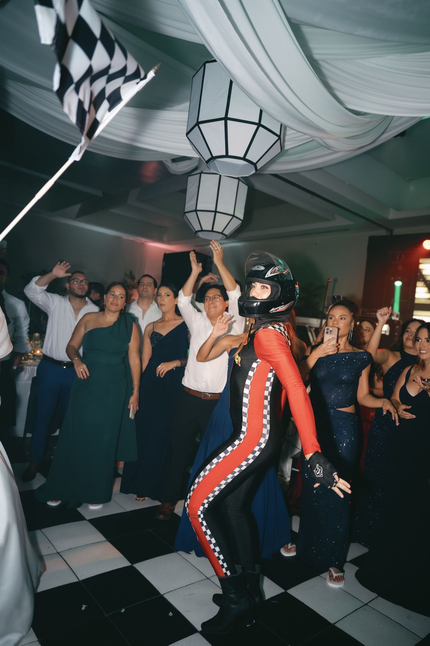 Energetic crowd dances and celebrates at a dimly lit indoor party with geometric ceiling lights.