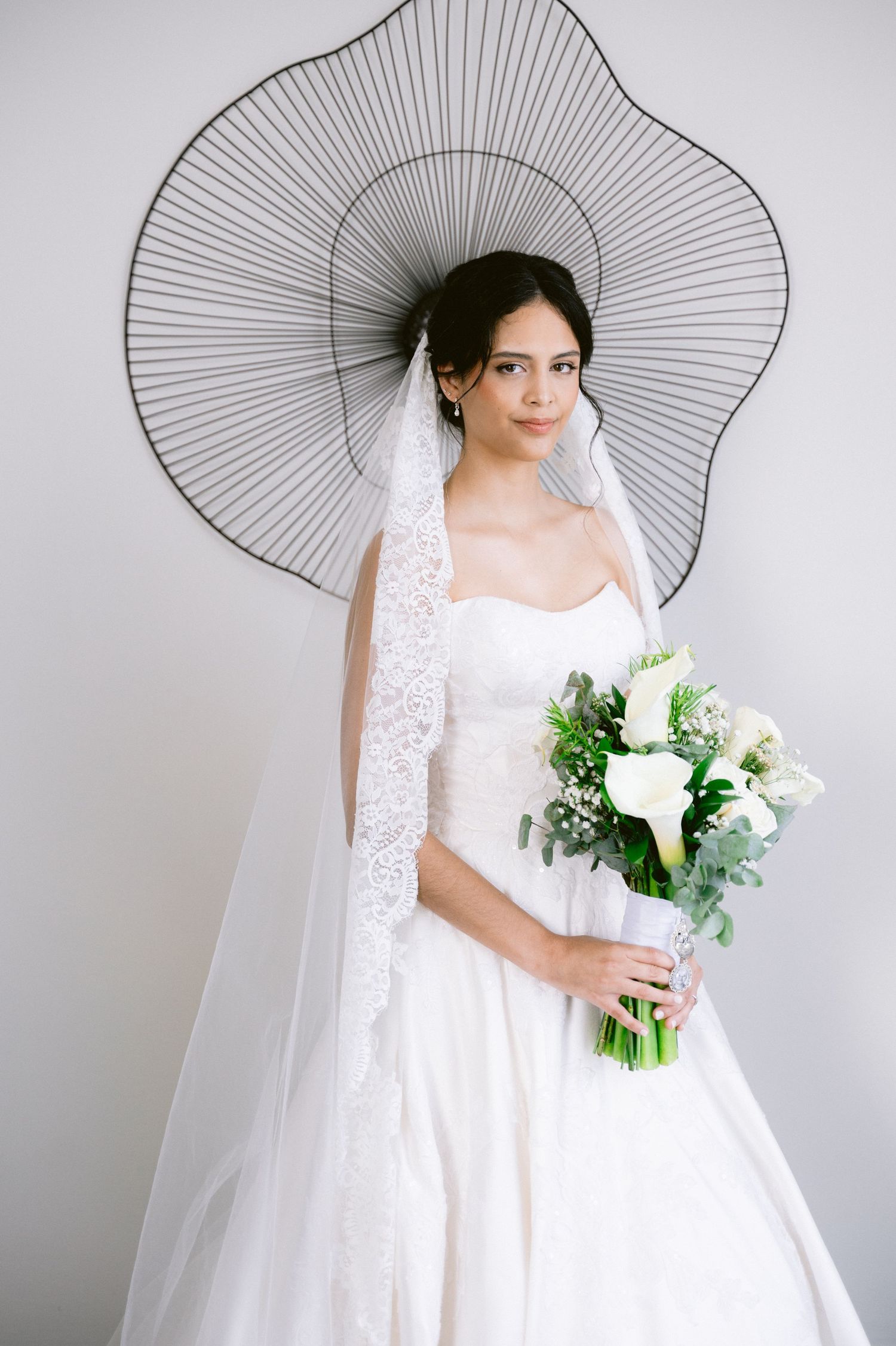 Bride in white lace dress poses with bouquet against geometric backdrop.