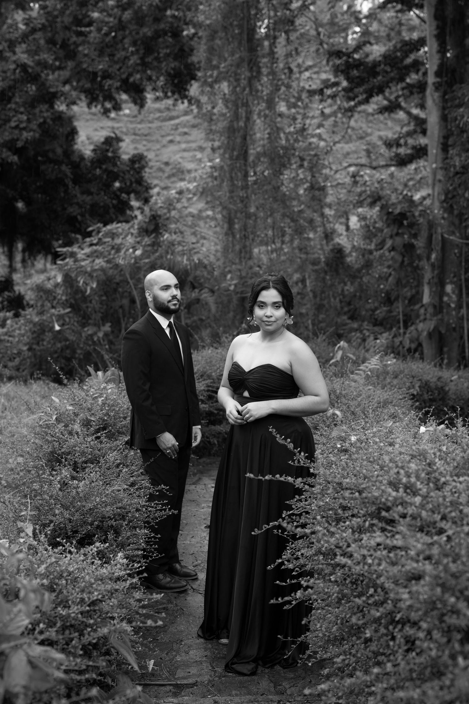 A couple in formal attire poses together in a wooded outdoor setting for romantic black and white photos.