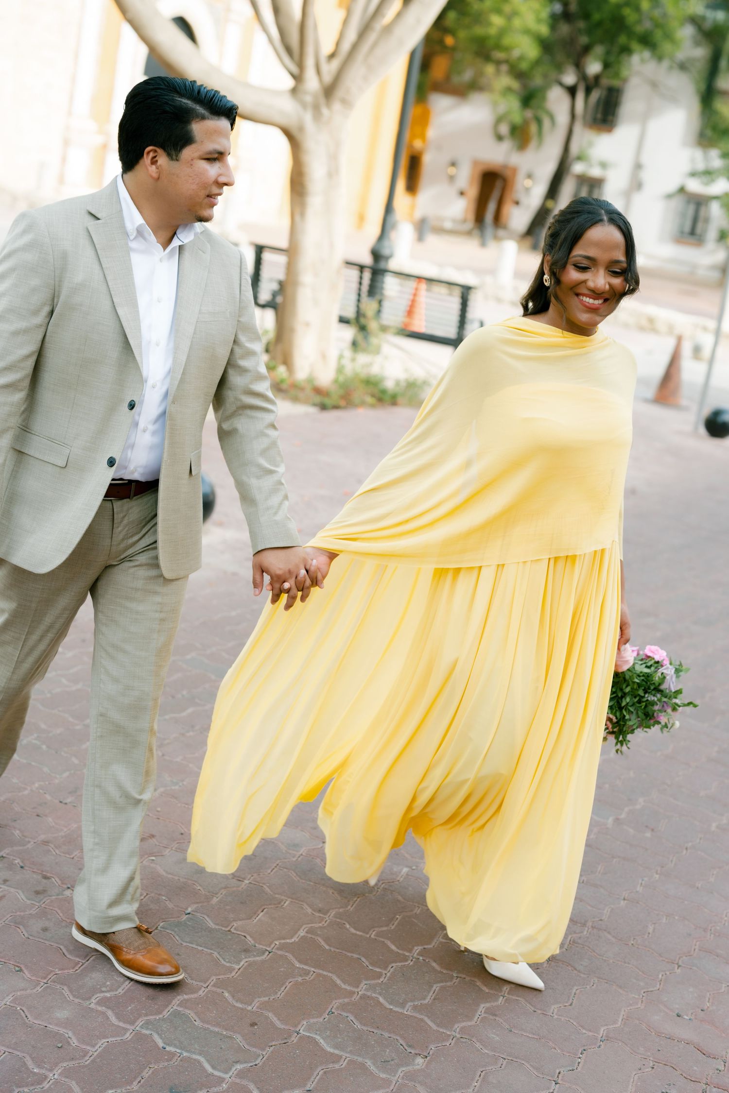 A couple walking together on a paved street, with one person wearing a yellow flowing dress and the other in a gray suit.