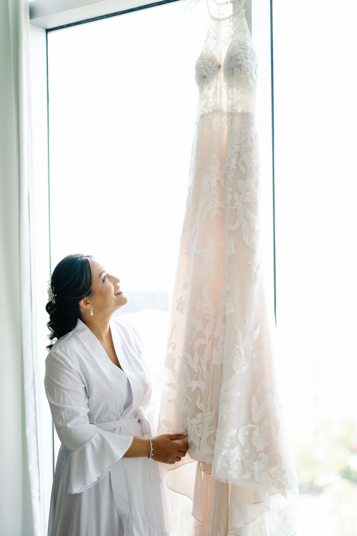 Woman in white robe admires a delicate lace wedding dress hanging by sunlit windows.