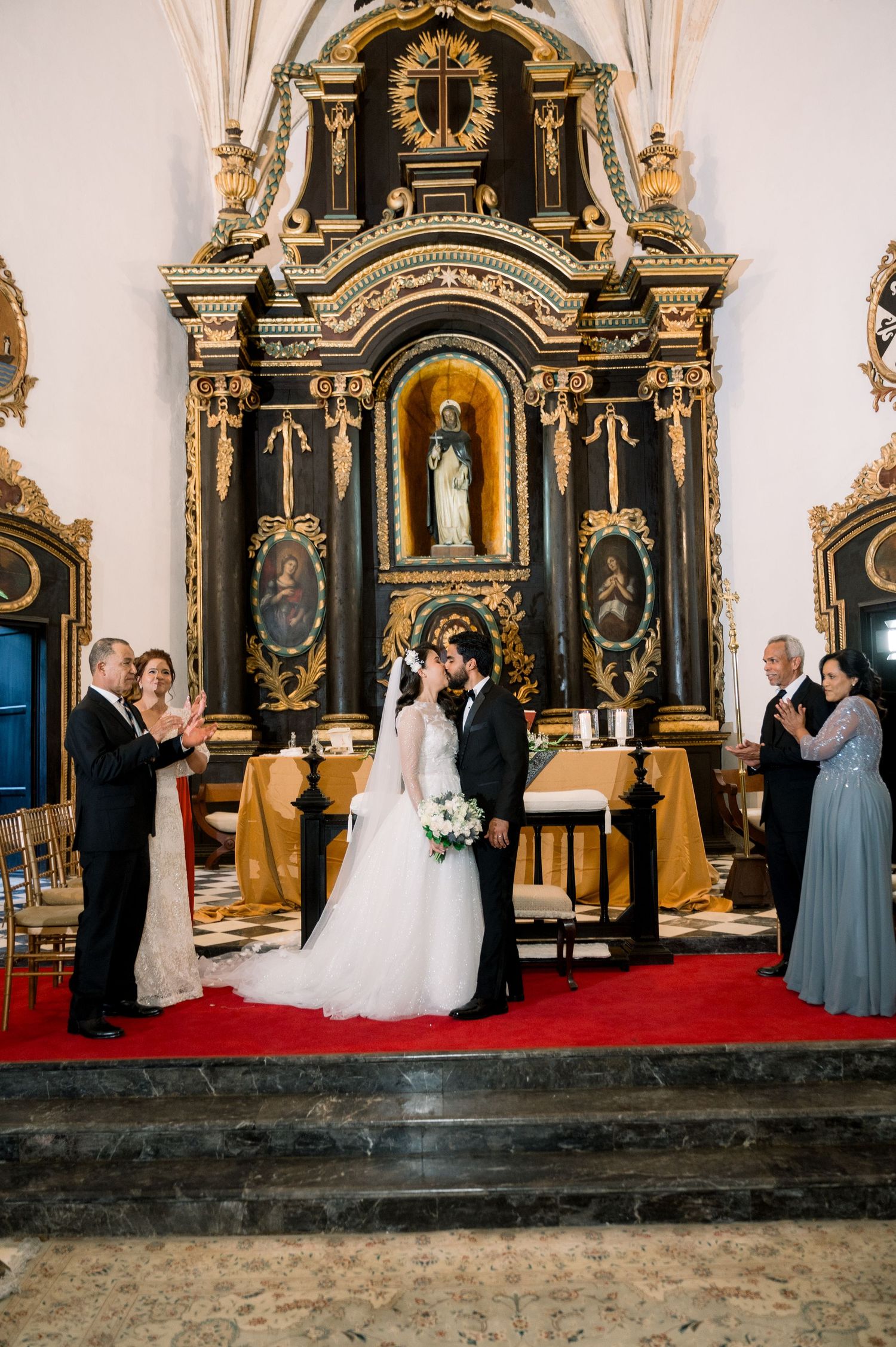 Wedding ceremony taking place in an ornate historic church with golden baroque altarpiece and red carpet.