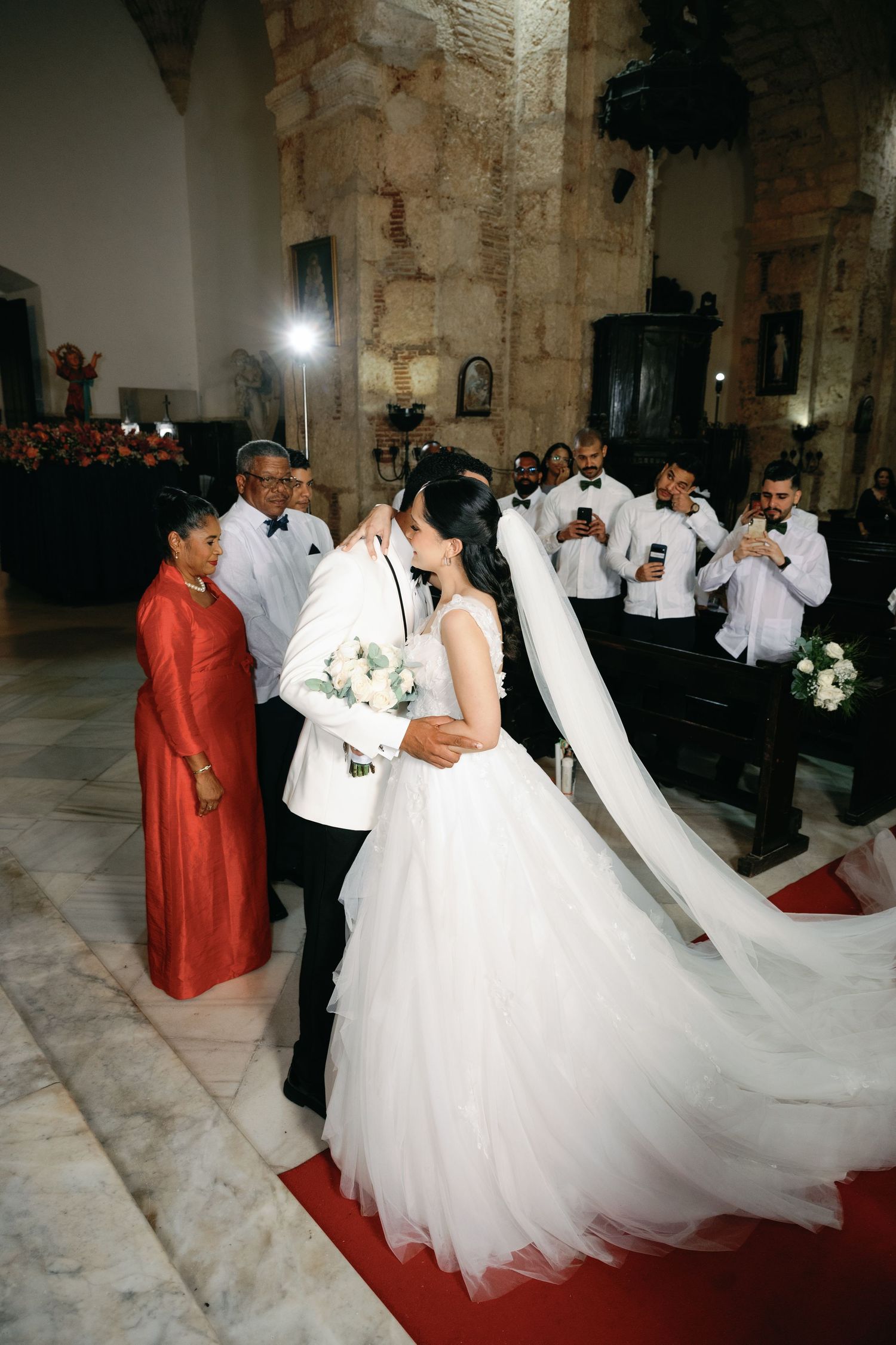 Multiple couples in formal wedding attire gather in an ornate church with white dresses and red carpet.