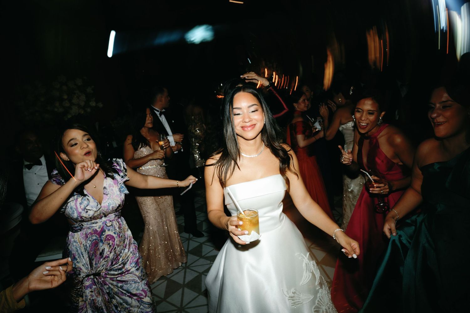 A bride in a white dress dances joyfully on a dark dance floor surrounded by celebrating wedding guests.