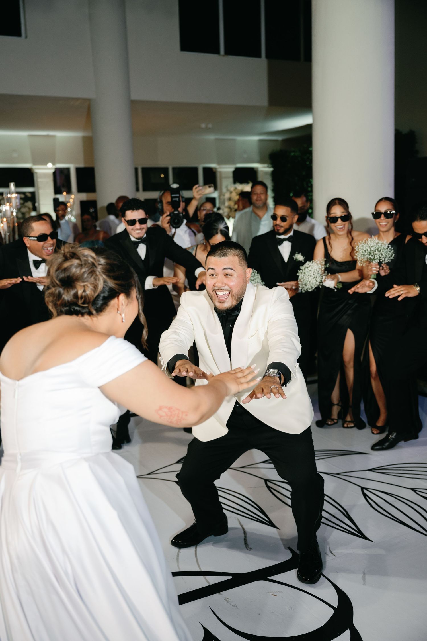 Wedding guests in black and white attire performing synchronized dance moves on patterned floor.