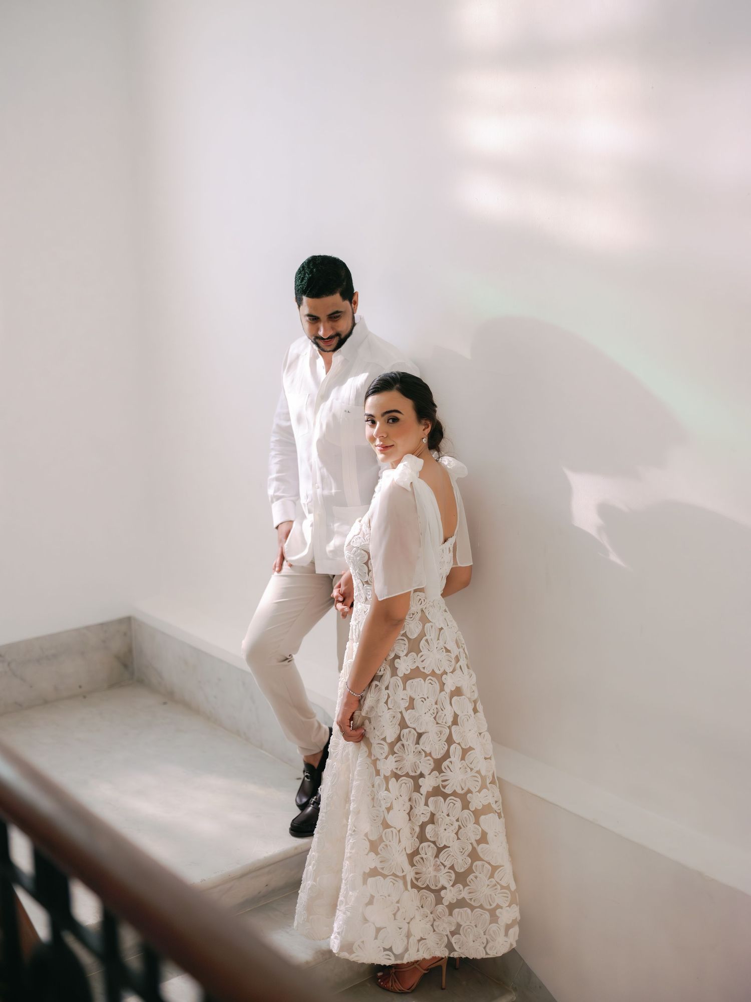 A couple in white attire poses together in a bright minimalist setting during their wedding photoshoot.