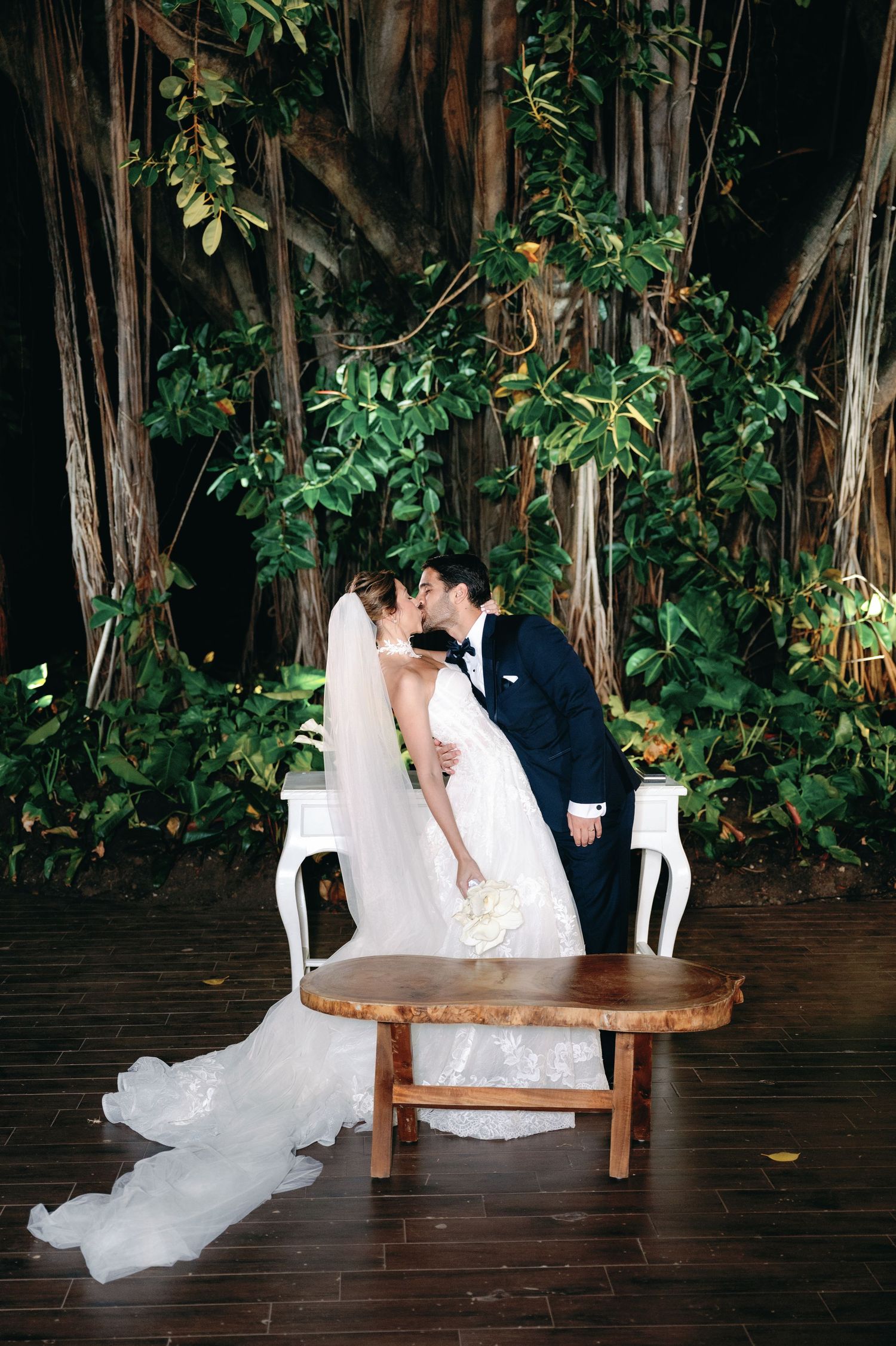 A romantic wedding couple shares an intimate moment on a wooden bench surrounded by lush garden foliage.