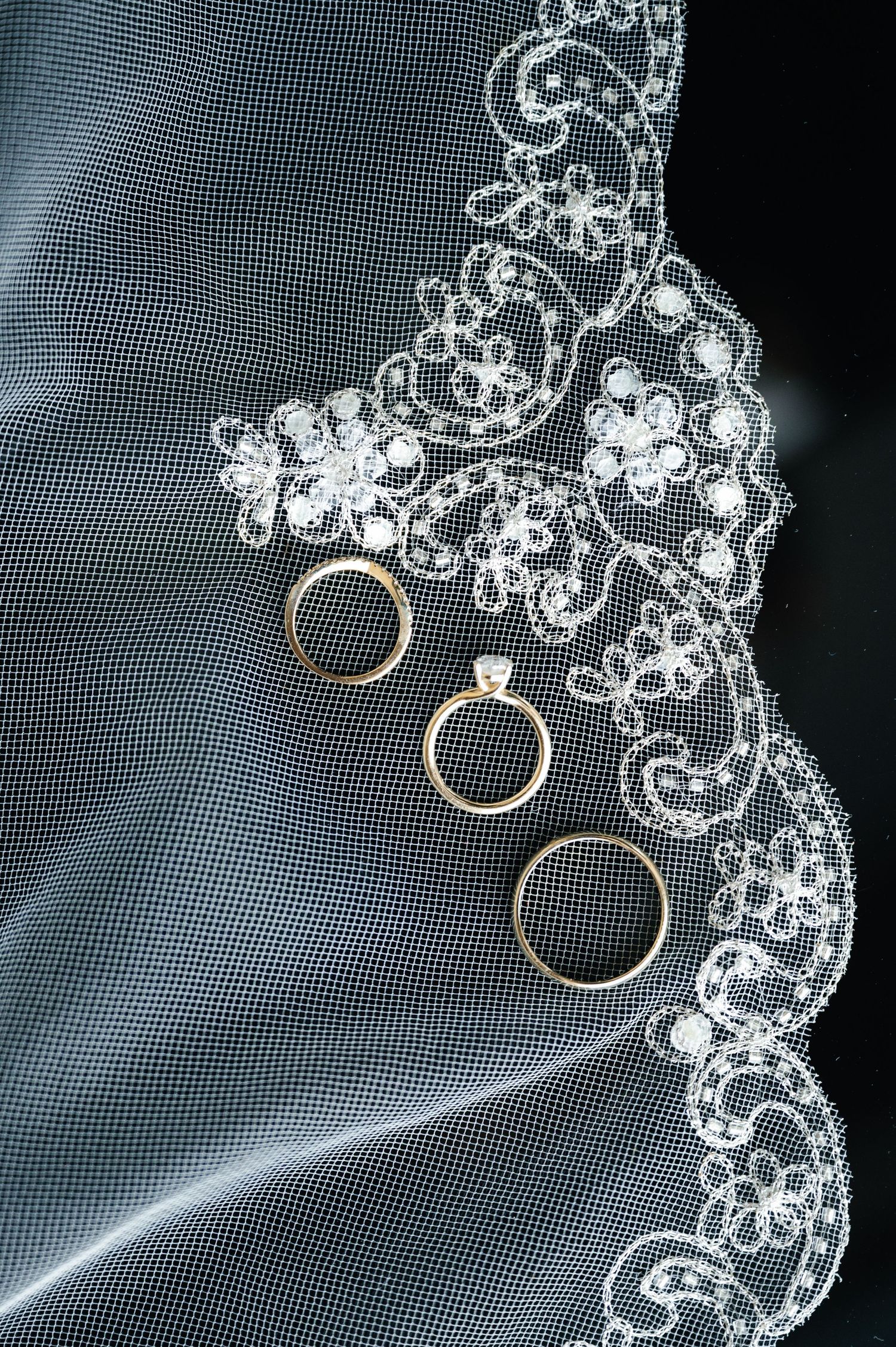 Three gold wedding rings arranged vertically on delicate white lace with floral embroidery against dark background.