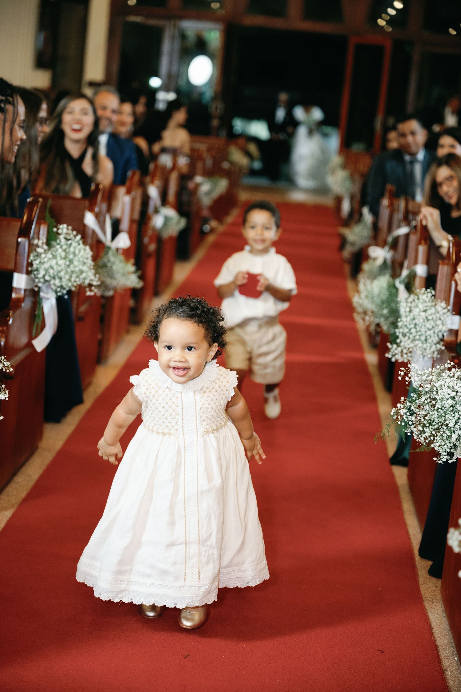 Young flower girl in white dress walks down red-carpeted church aisle decorated with baby's breath flowers.
