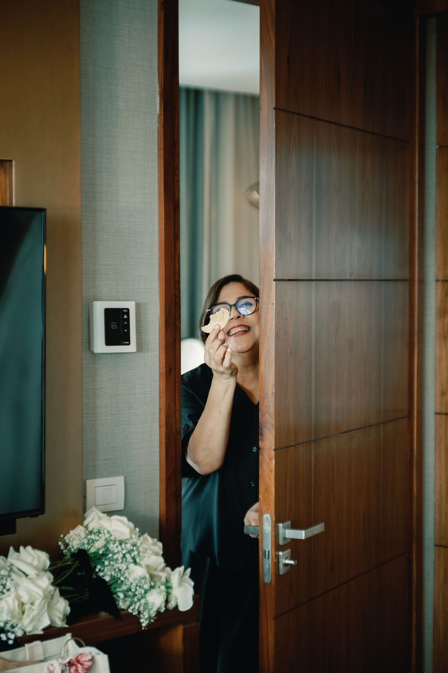 Someone peeks playfully from behind a modern wooden door with a digital thermostat mounted on the wall.