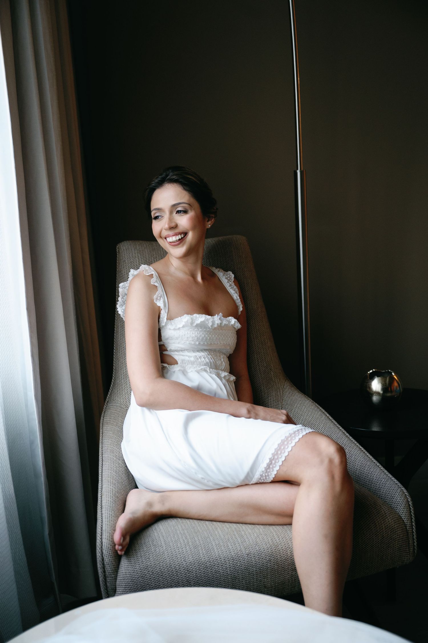 Woman in white dress lounges on gray chair in intimate boudoir photography series.