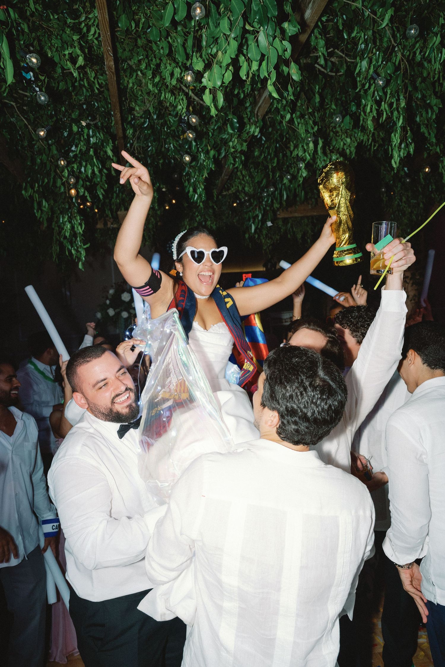 Celebratory party scene with World Cup trophy being held up among crowd under green foliage.
