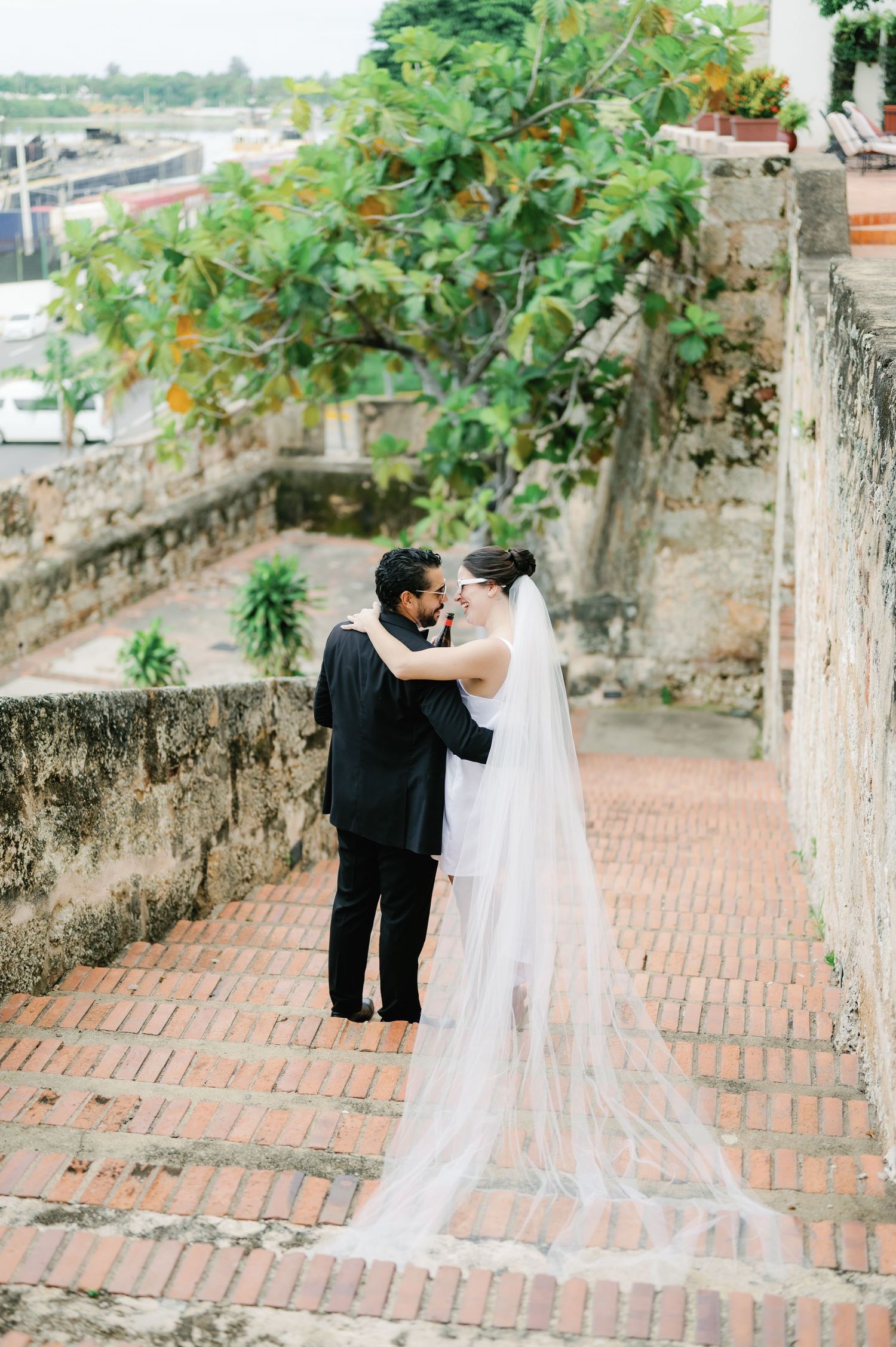 A couple in wedding attire embracing on brick steps with greenery and stone walls in the background.