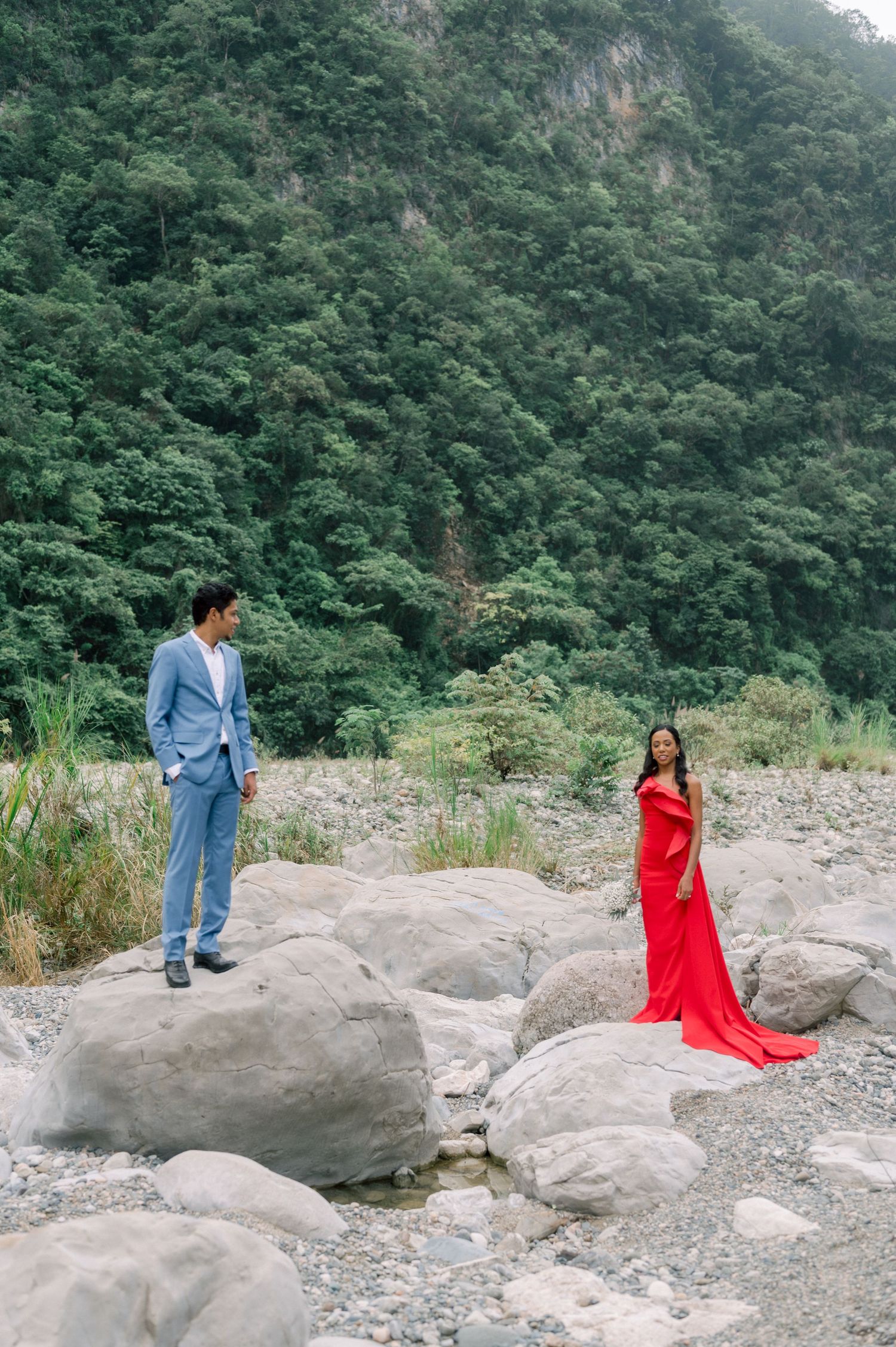 Two people in contrasting blue and red outfits pose on rocky terrain against a lush mountain backdrop.