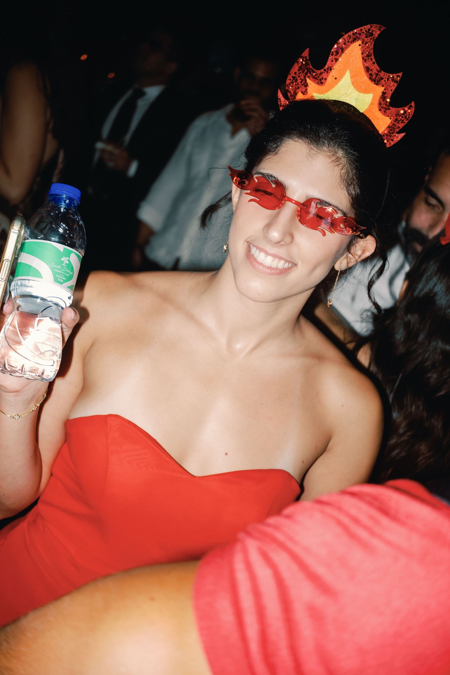 A partygoer in a red dress and flame crown accessory holds a water bottle while smiling at a nighttime event.