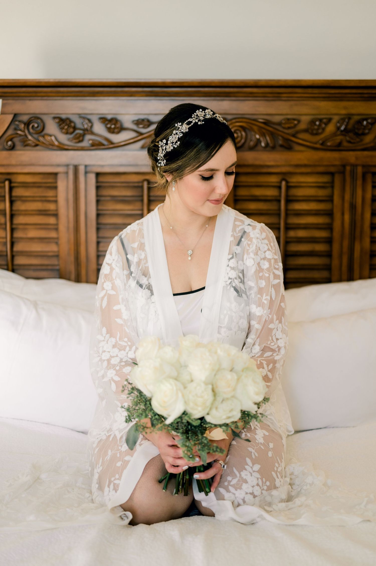 A bride in white lace holding a bouquet of white roses while sitting on a bed with wooden headboard.