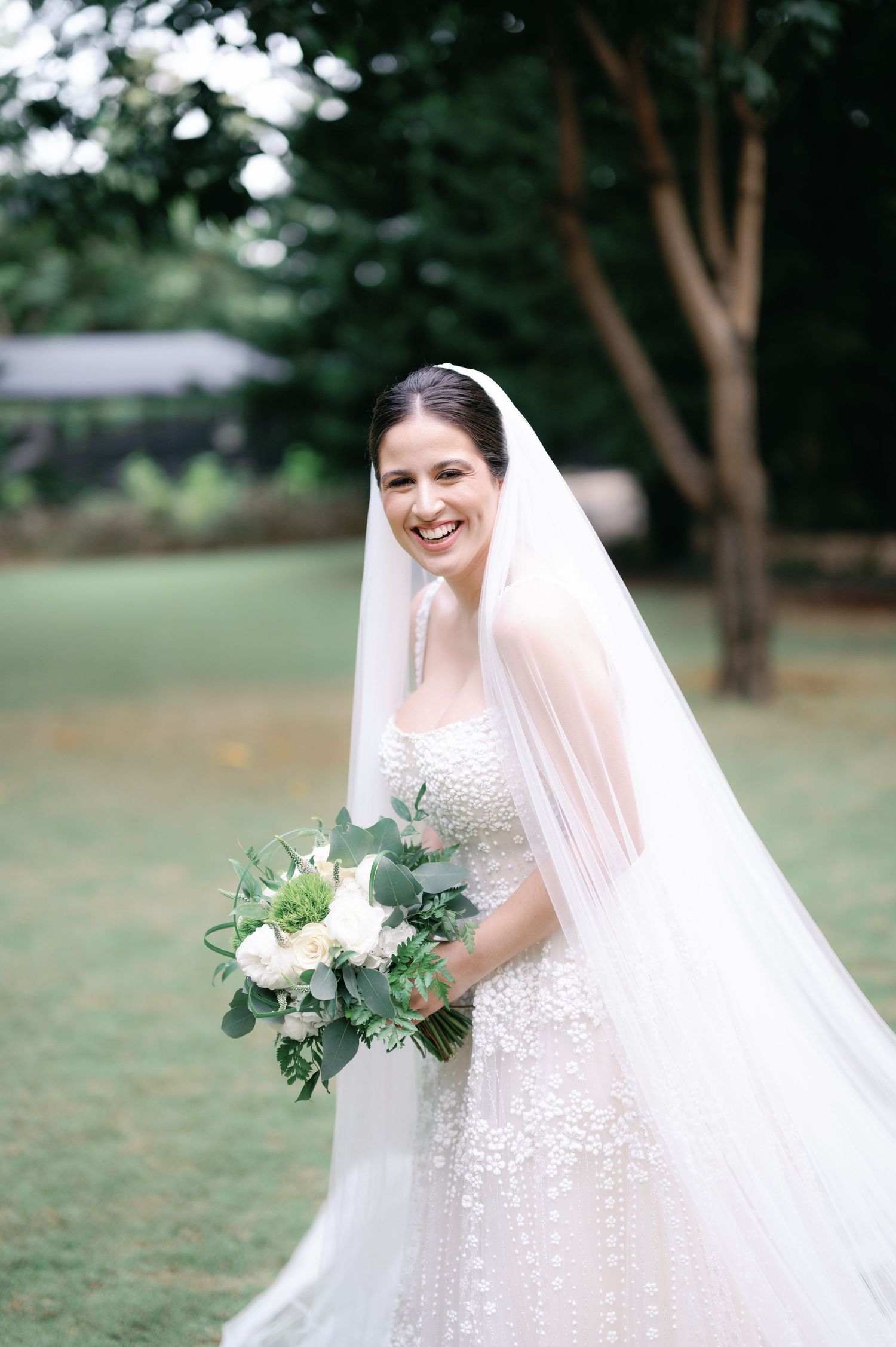 A bride in a detailed lace wedding gown holds a white and green bouquet in an outdoor garden setting.