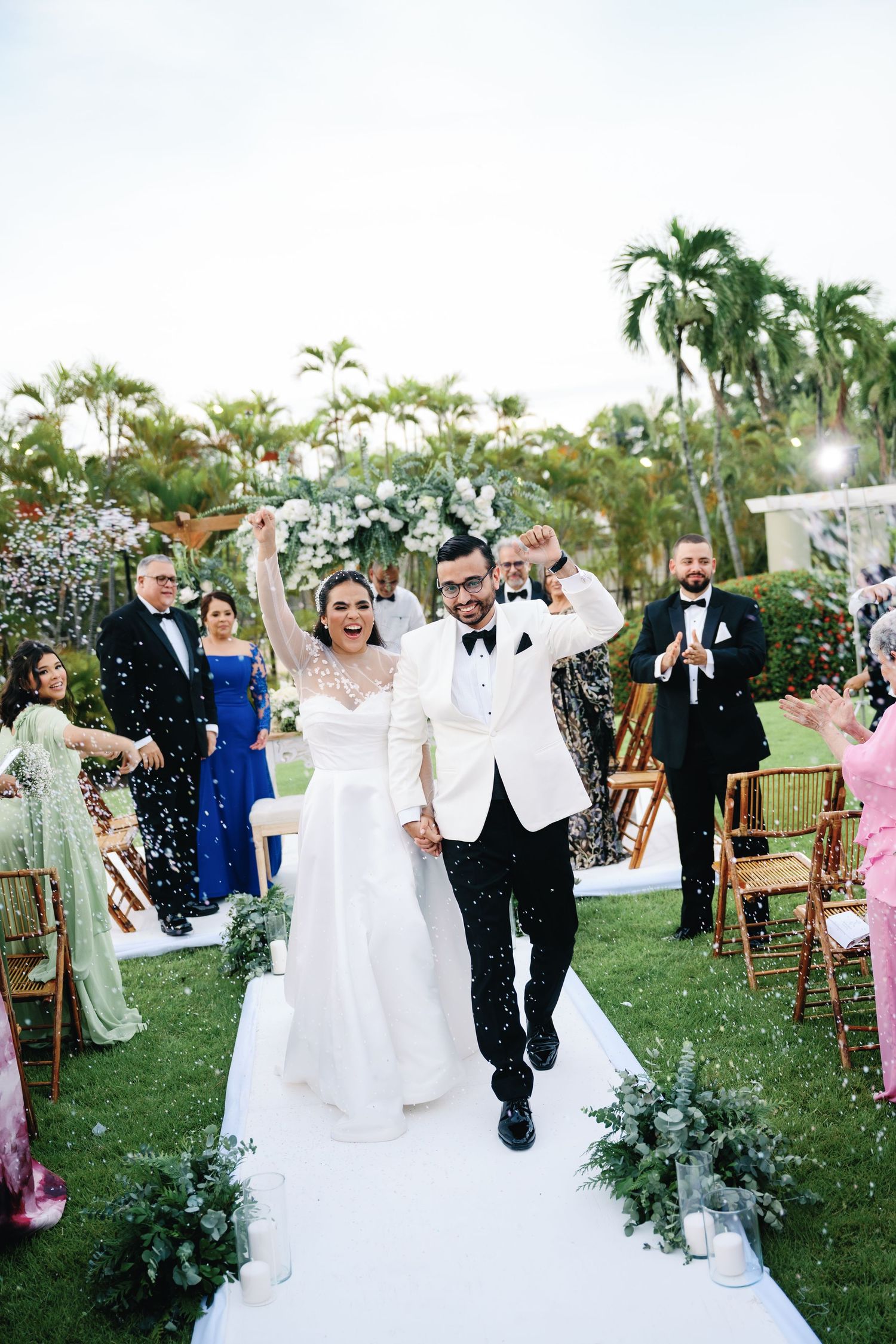 Joyful celebration moment at an outdoor wedding with guests throwing confetti on a white aisle runner.