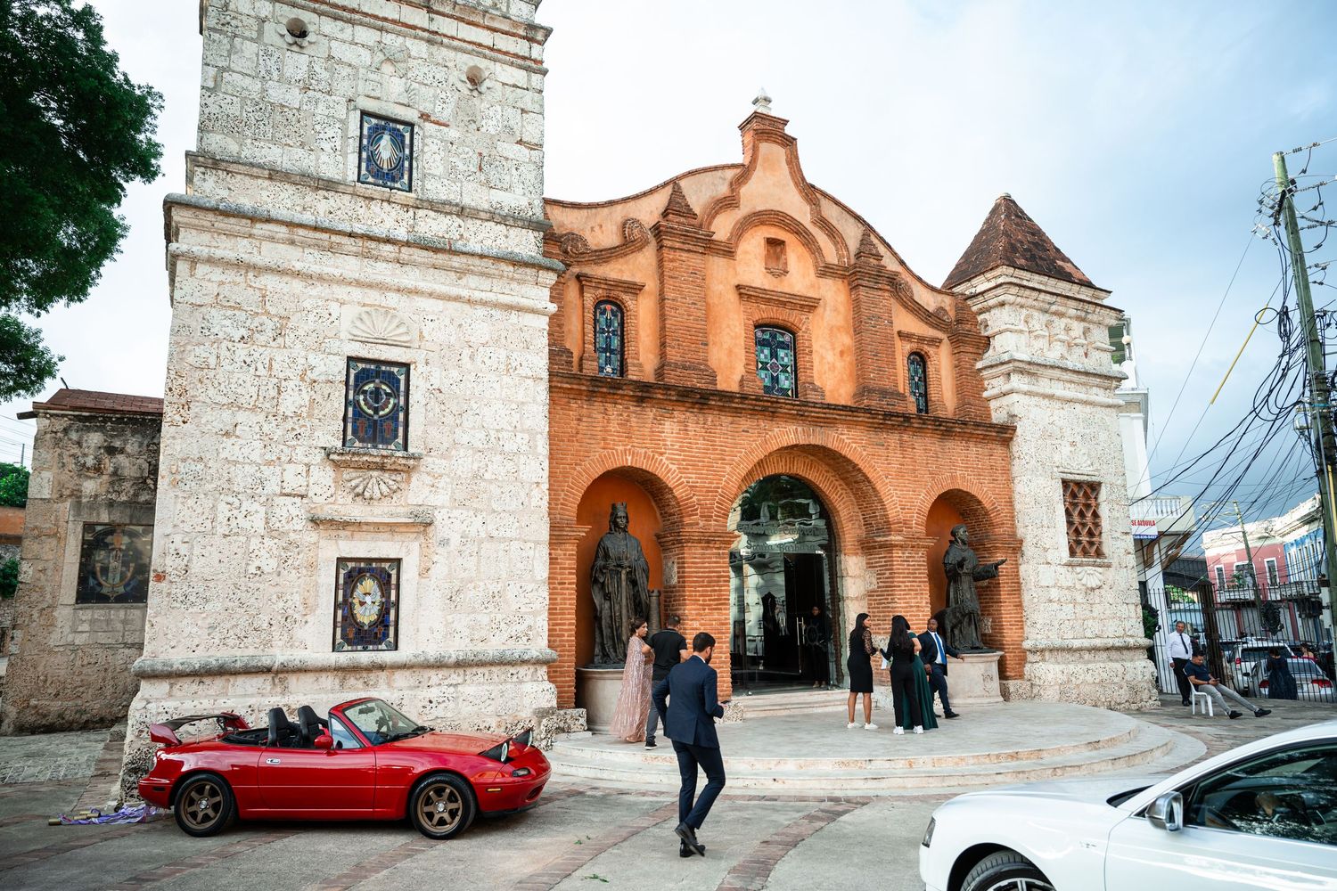 Historic stone church facade with red brick accents and pedestrians walking near parked cars on a city street.