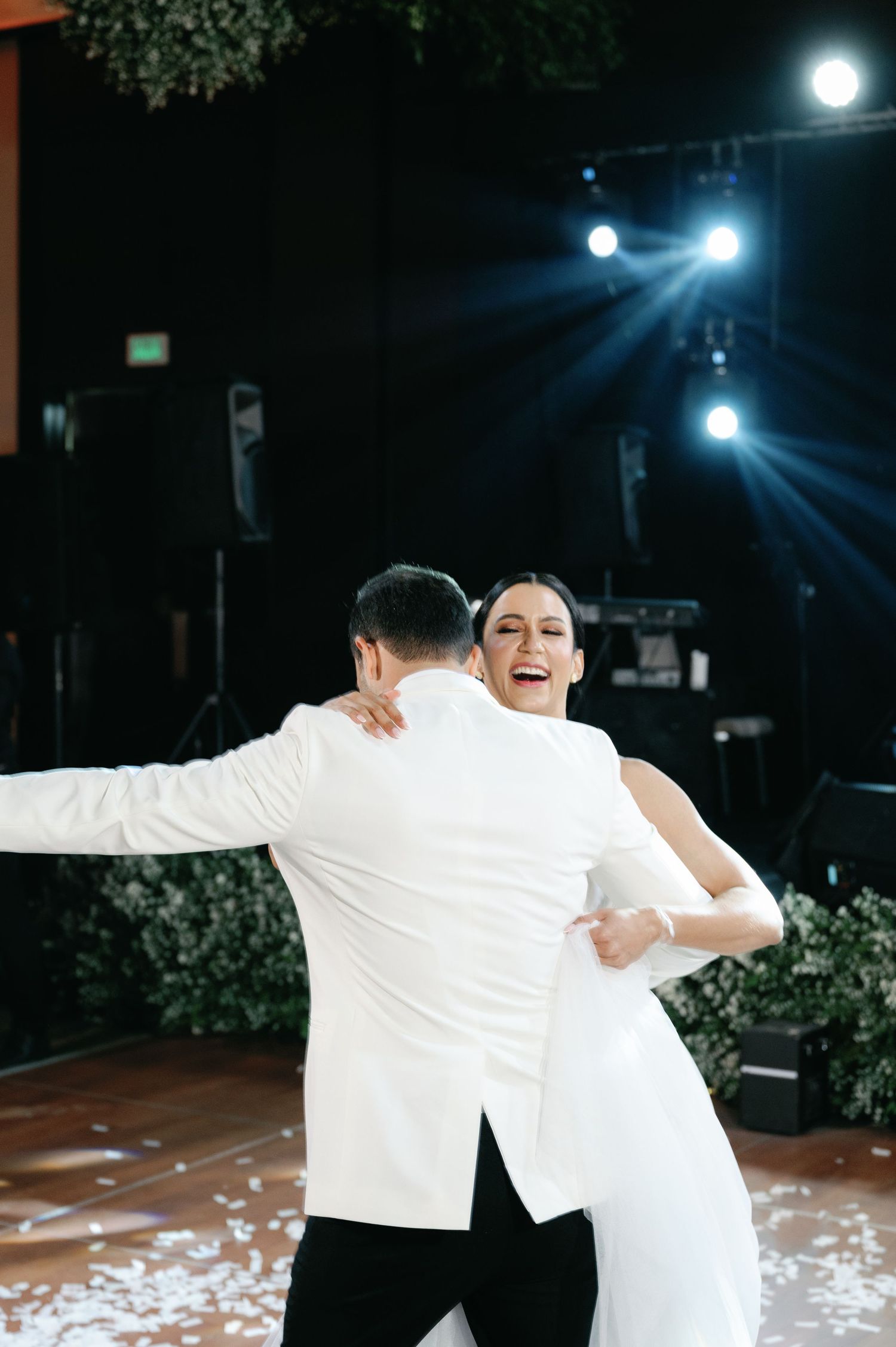 A couple shares a joyful dance moment outdoors at night under bright lights and surrounded by foliage.