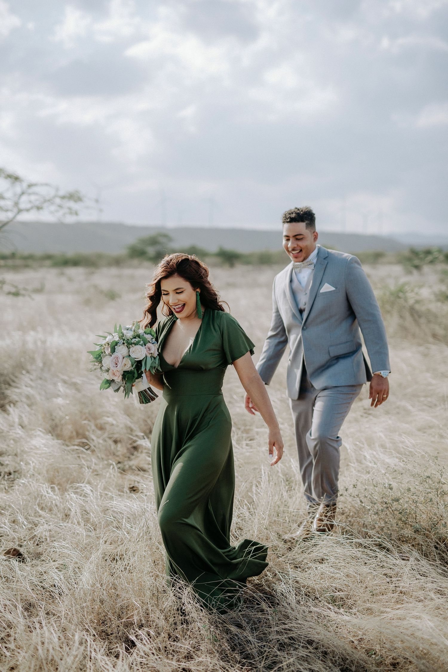 A couple walks playfully through a field of tall dried grass on an overcast day.