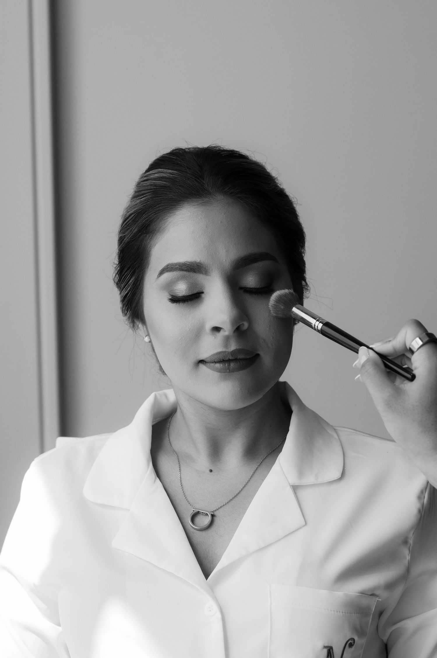 A makeup artist applies cosmetics to someone wearing a white collared top in a black and white photo series.