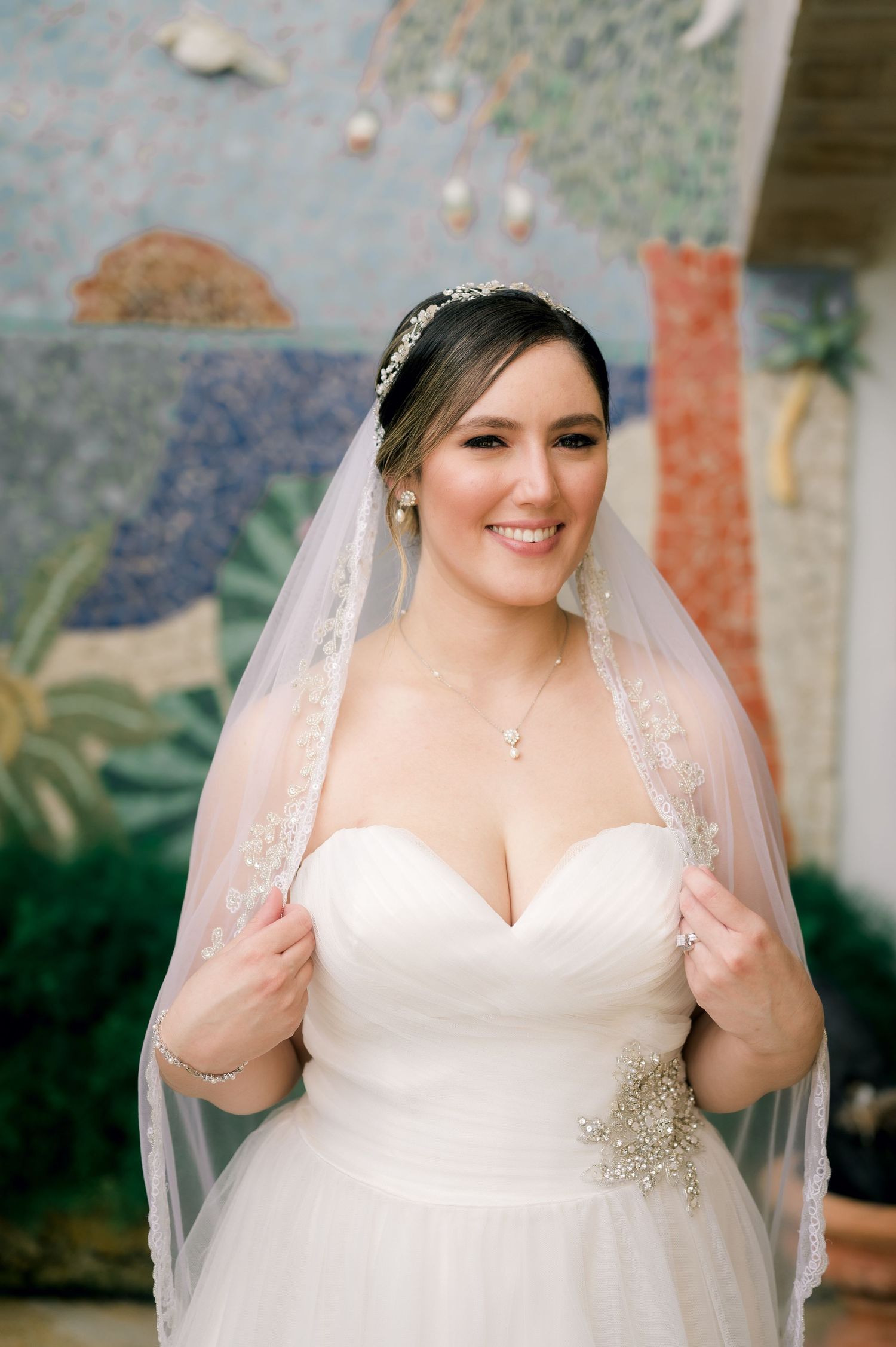 A bride in a classic white wedding gown adjusts her veil while posing outdoors.