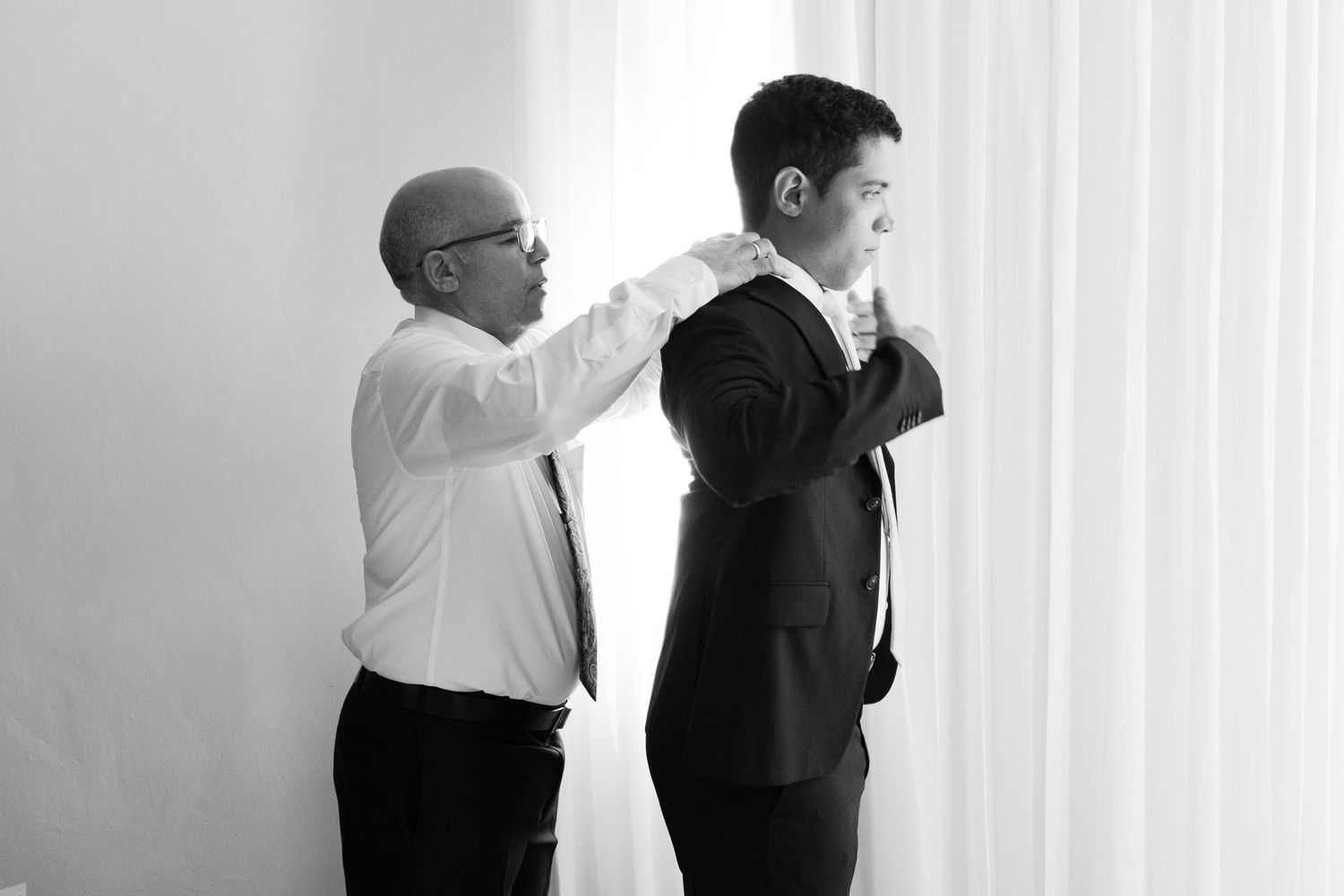 A man helps adjust another man's suit jacket while standing against a white wall.