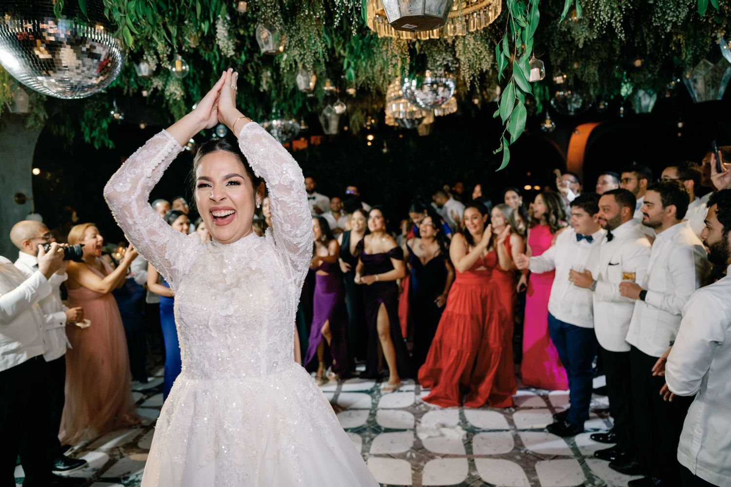 Wedding celebration on a dance floor with guests dancing under greenery and hanging decorations at an indoor venue.