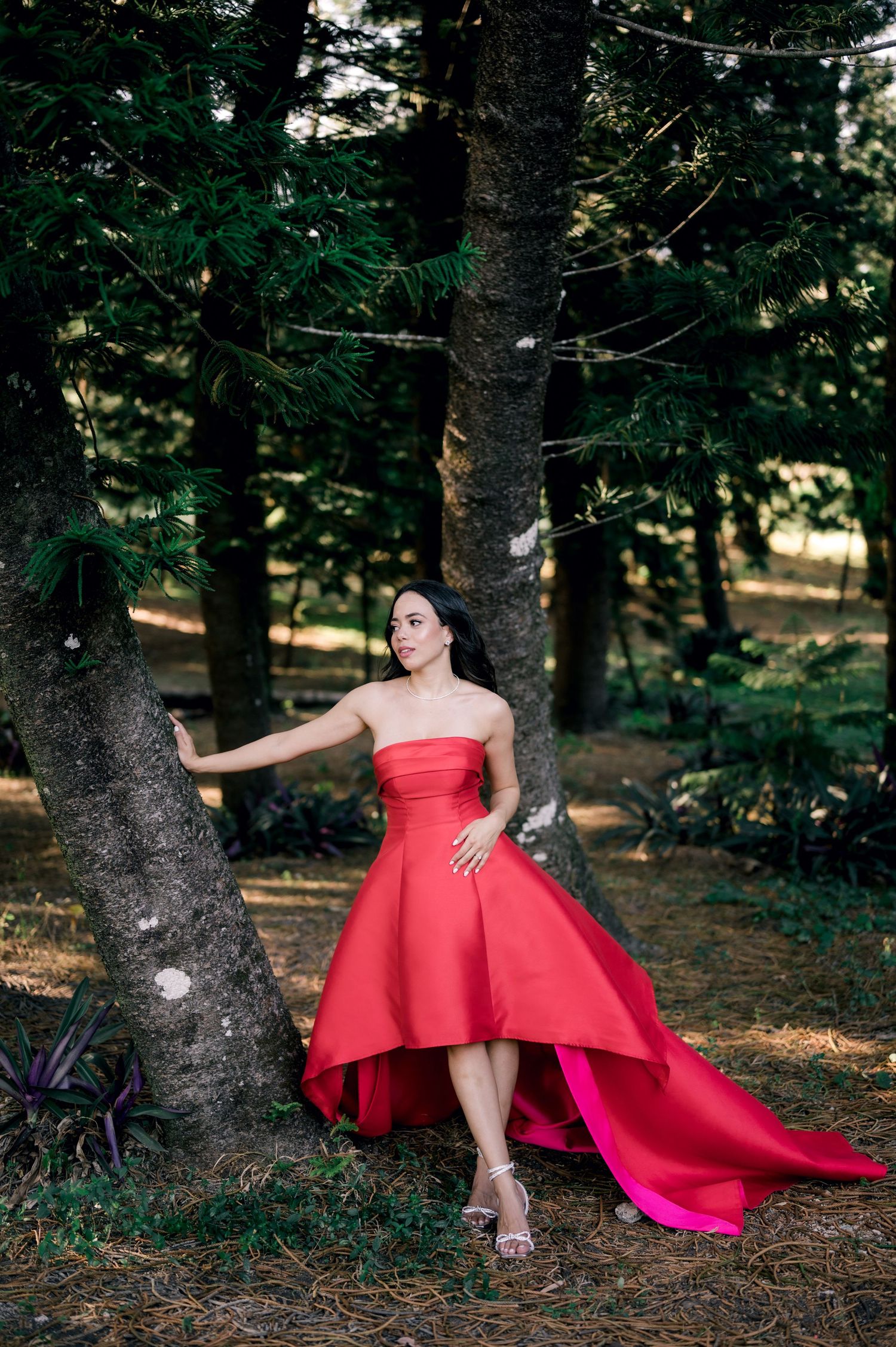 A person in a flowing red formal gown poses elegantly against a backdrop of tall trees in a forest setting.