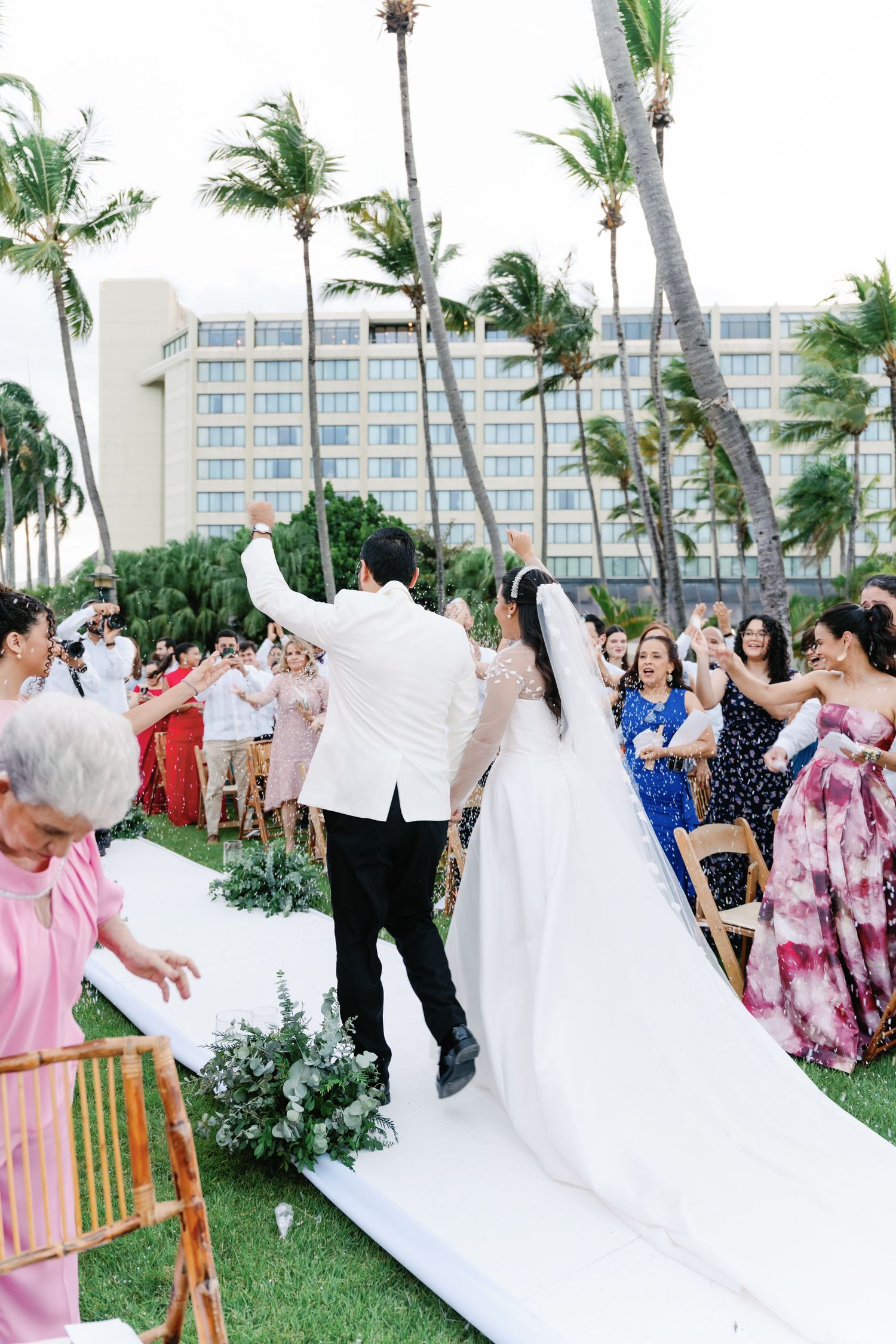 Wedding celebration with guests cheering as couple walks down white aisle under palm trees.