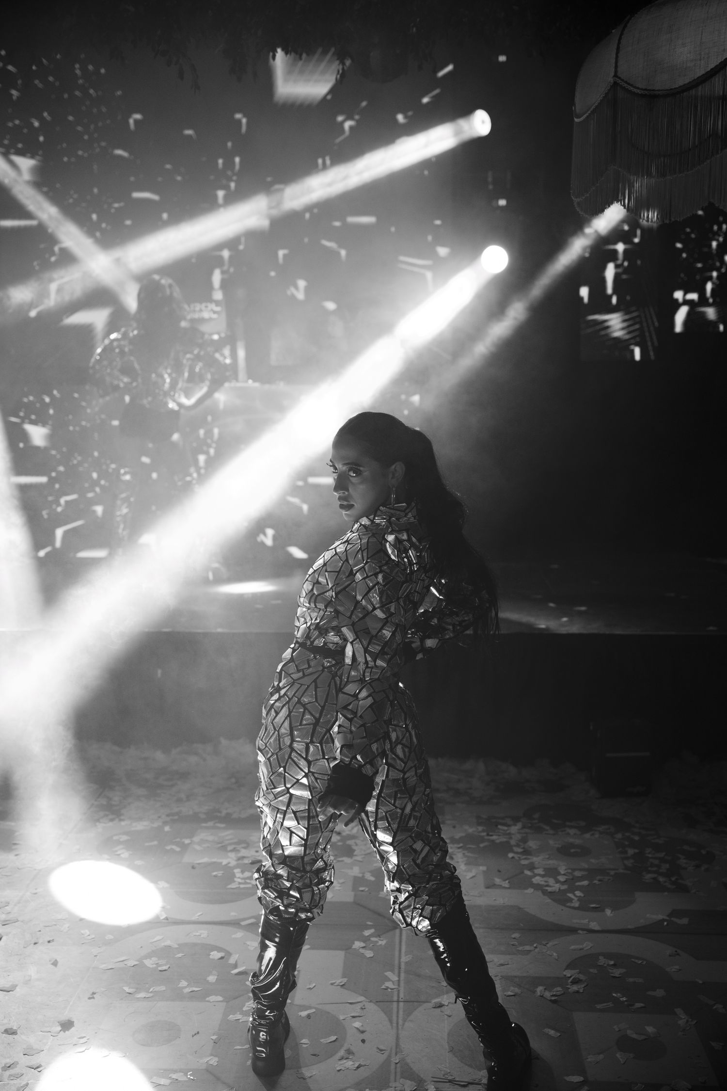 A performer in reflective gear dances under dramatic spotlights on a dark stage with falling confetti.