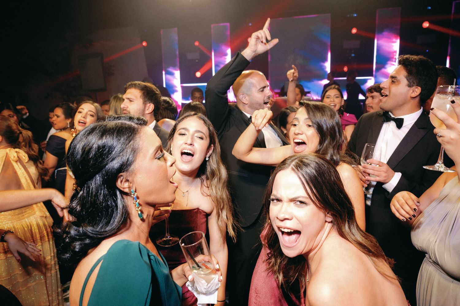 Excited crowd of formal event attendees dancing and singing together under colorful lighting.