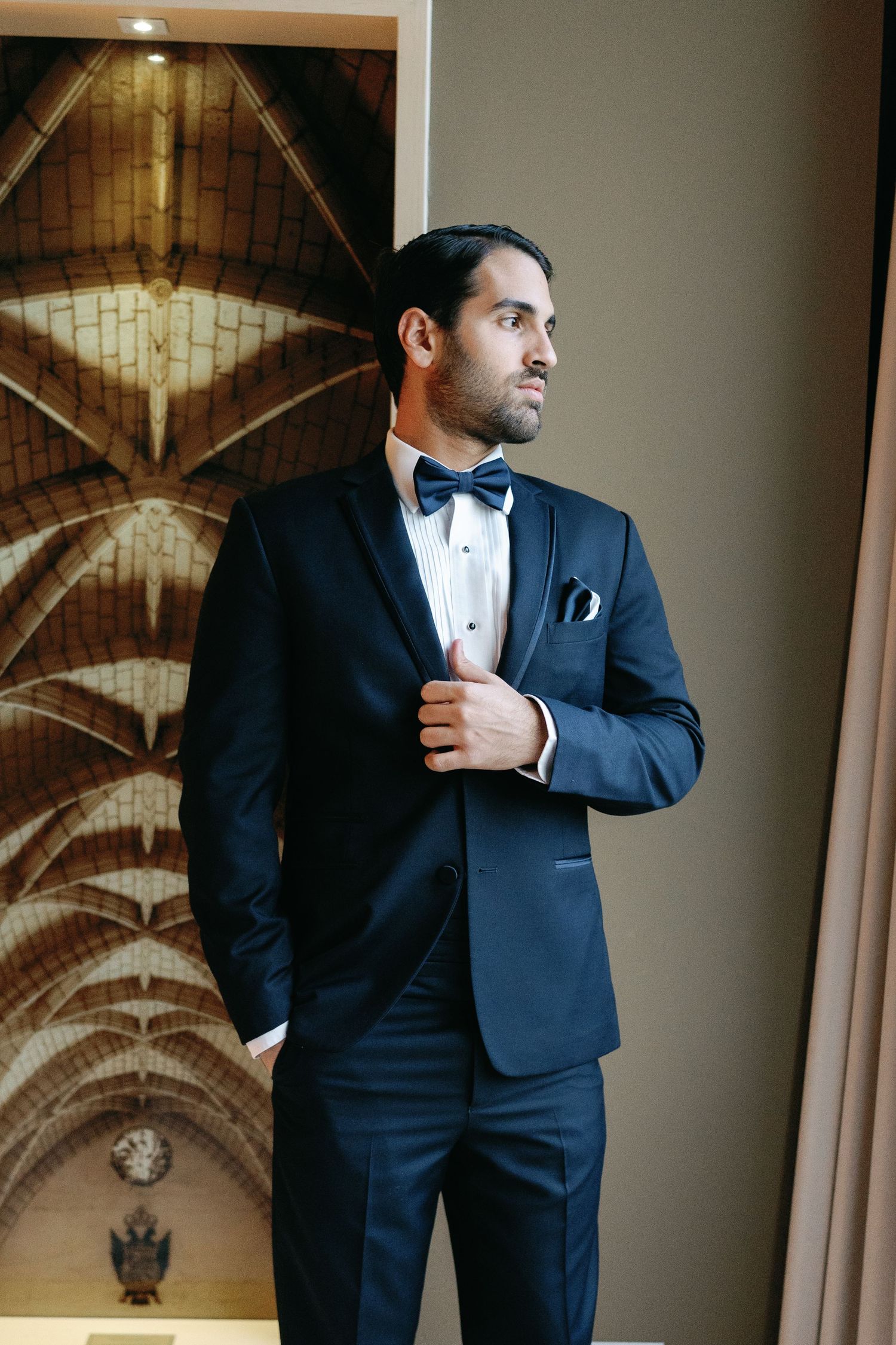 Stylish groom in classic black tuxedo posing against architectural backdrop in dramatic lighting.