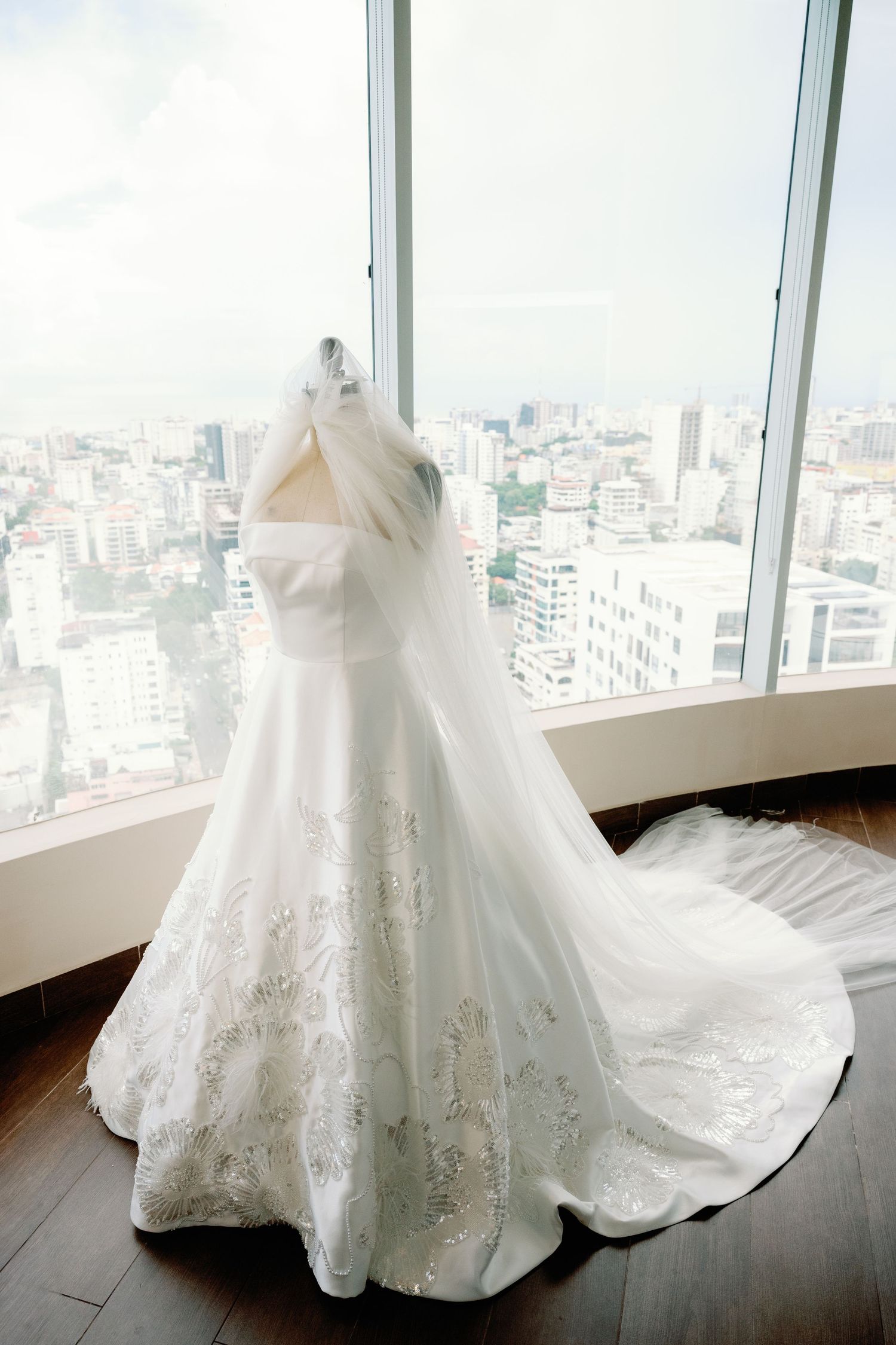 Elegant white wedding dress with lace details displayed against a cityscape backdrop through large windows.