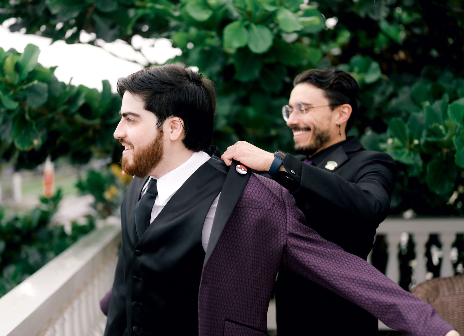 Groomsmen in dark suits laughing and celebrating together outdoors with natural greenery in background.