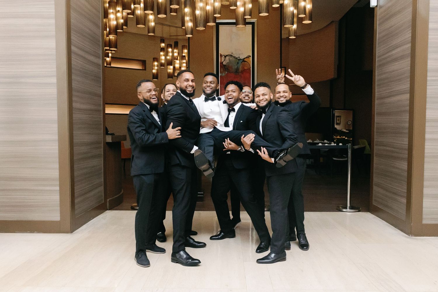 Wedding party members in formal black suits pose together on marble steps beneath warm lighting fixtures.