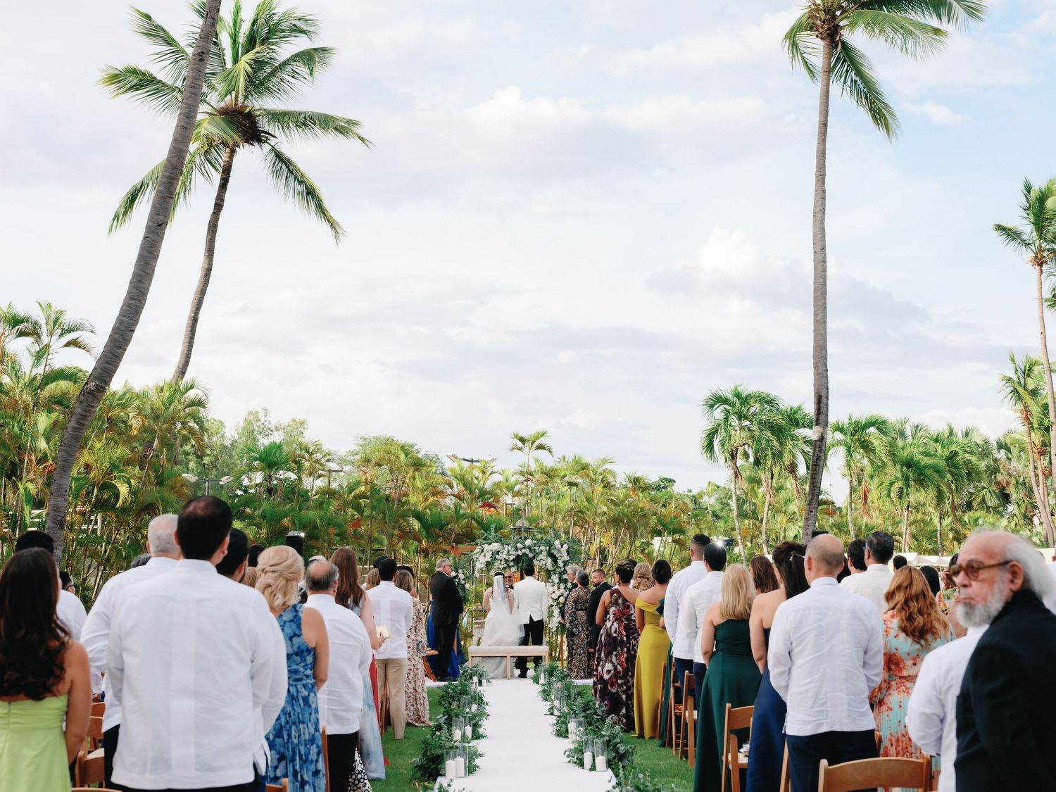 Outdoor wedding ceremony setup with palm trees and guests gathered under a tropical sky.