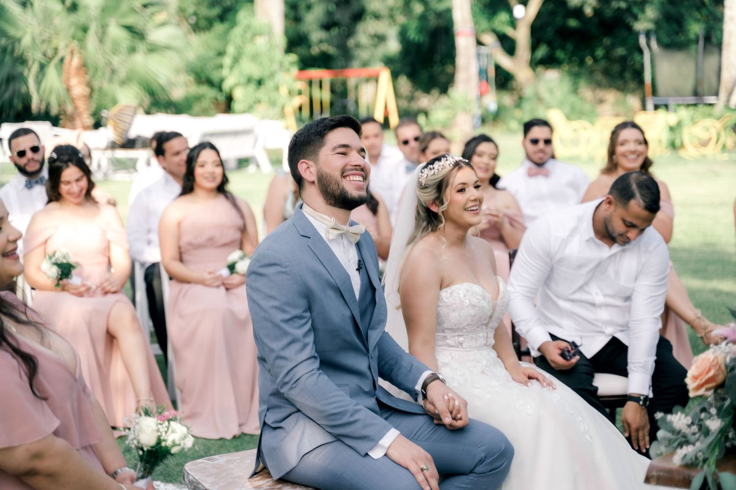 Wedding party in blush pink dresses and gray suits laugh together while sitting outdoors during summer celebration.