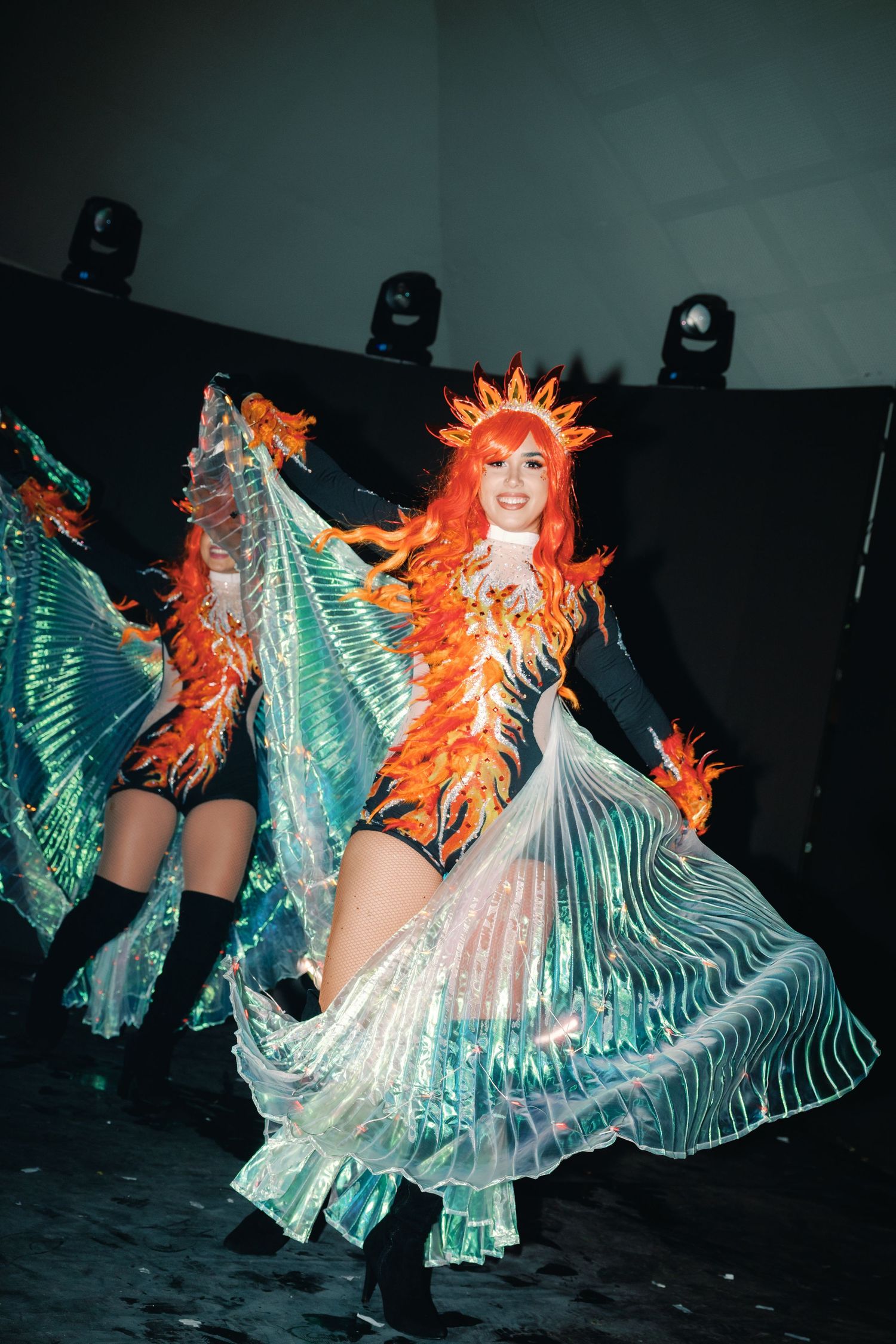 Dancers in shimmering turquoise wings and bright orange wigs perform a synchronized routine on a dark stage.