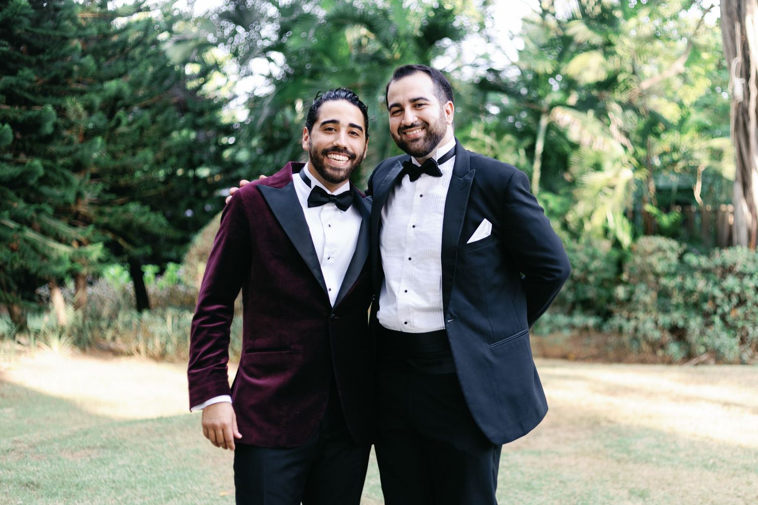 Two people in formal attire stand together smiling and laughing outdoors with trees in the background.