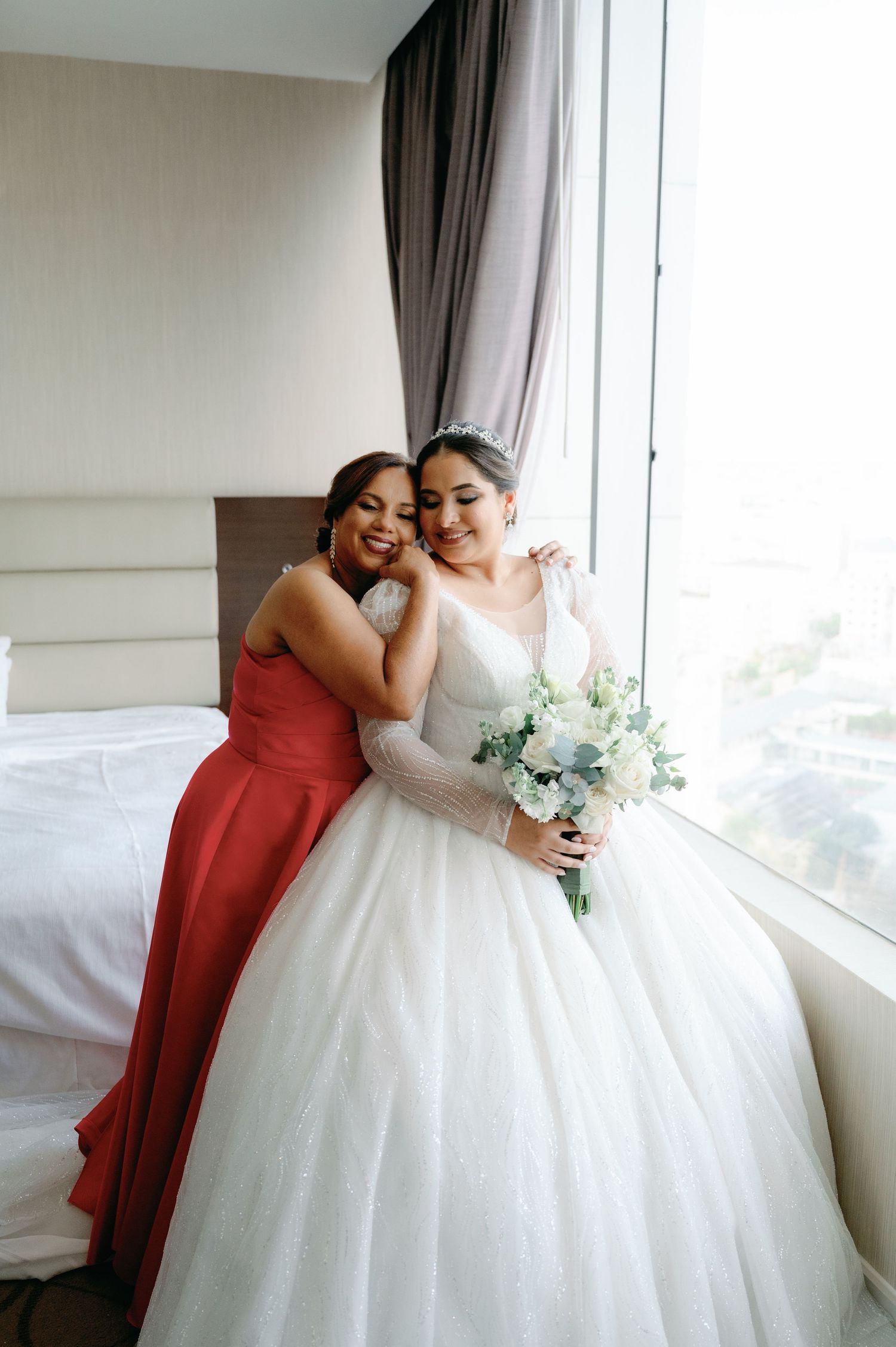 A bride in a white ballgown and bridesmaid in red share joyful moments near a bright window.
