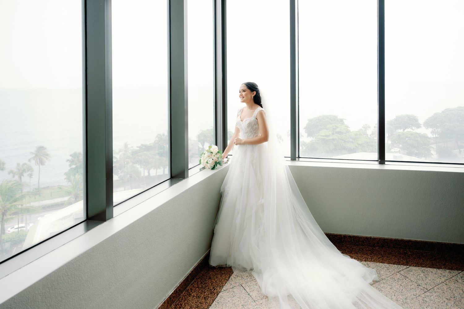 Bride in a flowing white wedding gown stands by large windows overlooking a city view in an elegant venue.