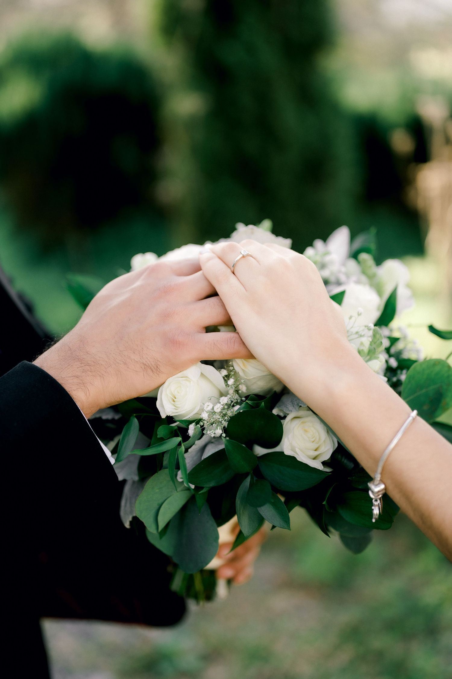 Two hands wearing wedding rings touch tenderly over white roses and greenery in a romantic gesture.