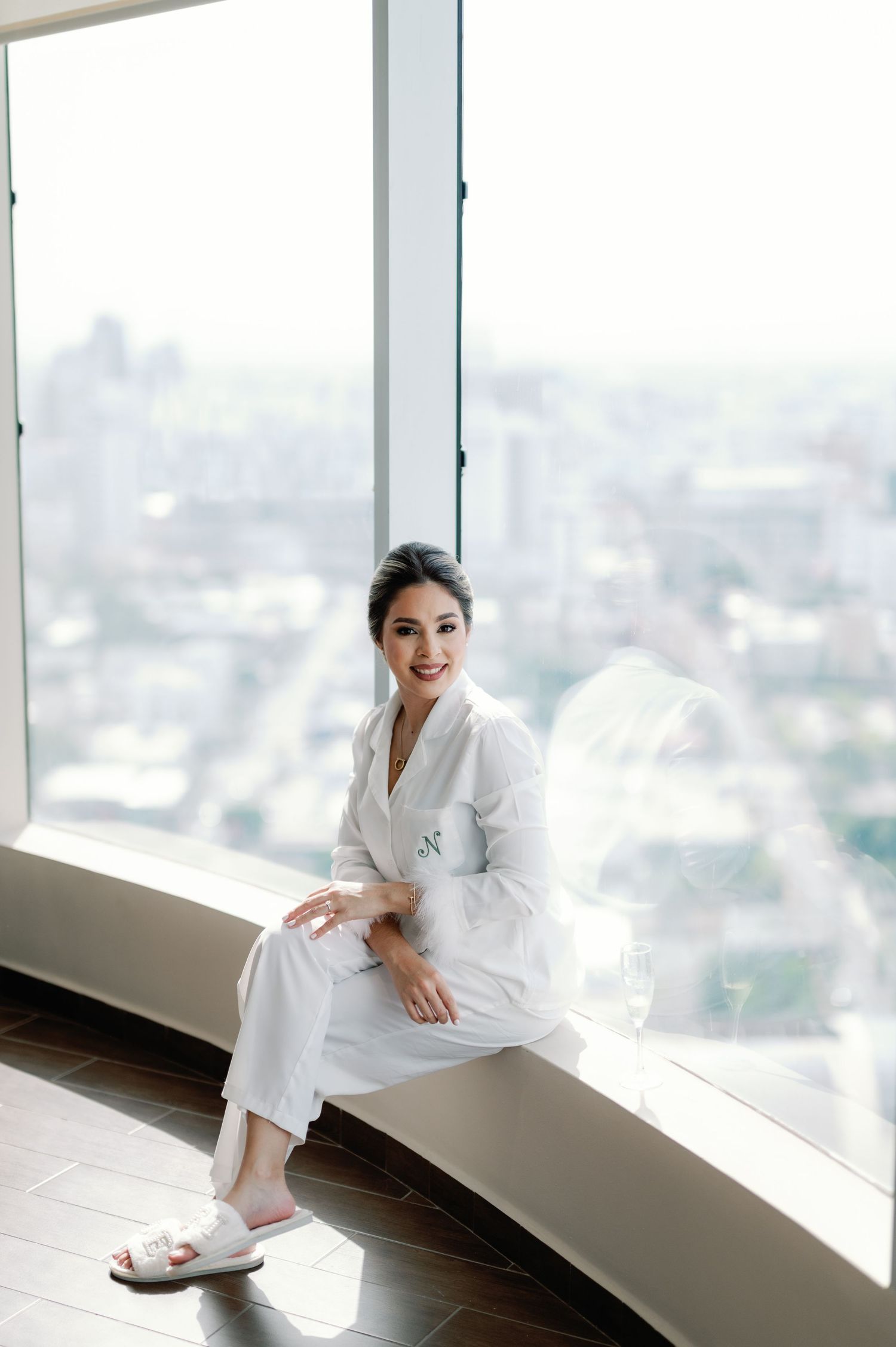 A person in a white robe relaxes by floor-to-ceiling windows in a high-rise building with city views.