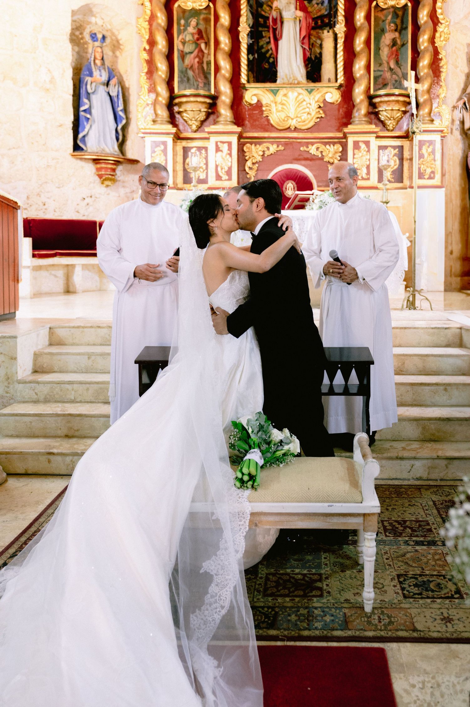 A wedding ceremony takes place at an ornate church altar with clergy in white robes and decorative religious artwork.