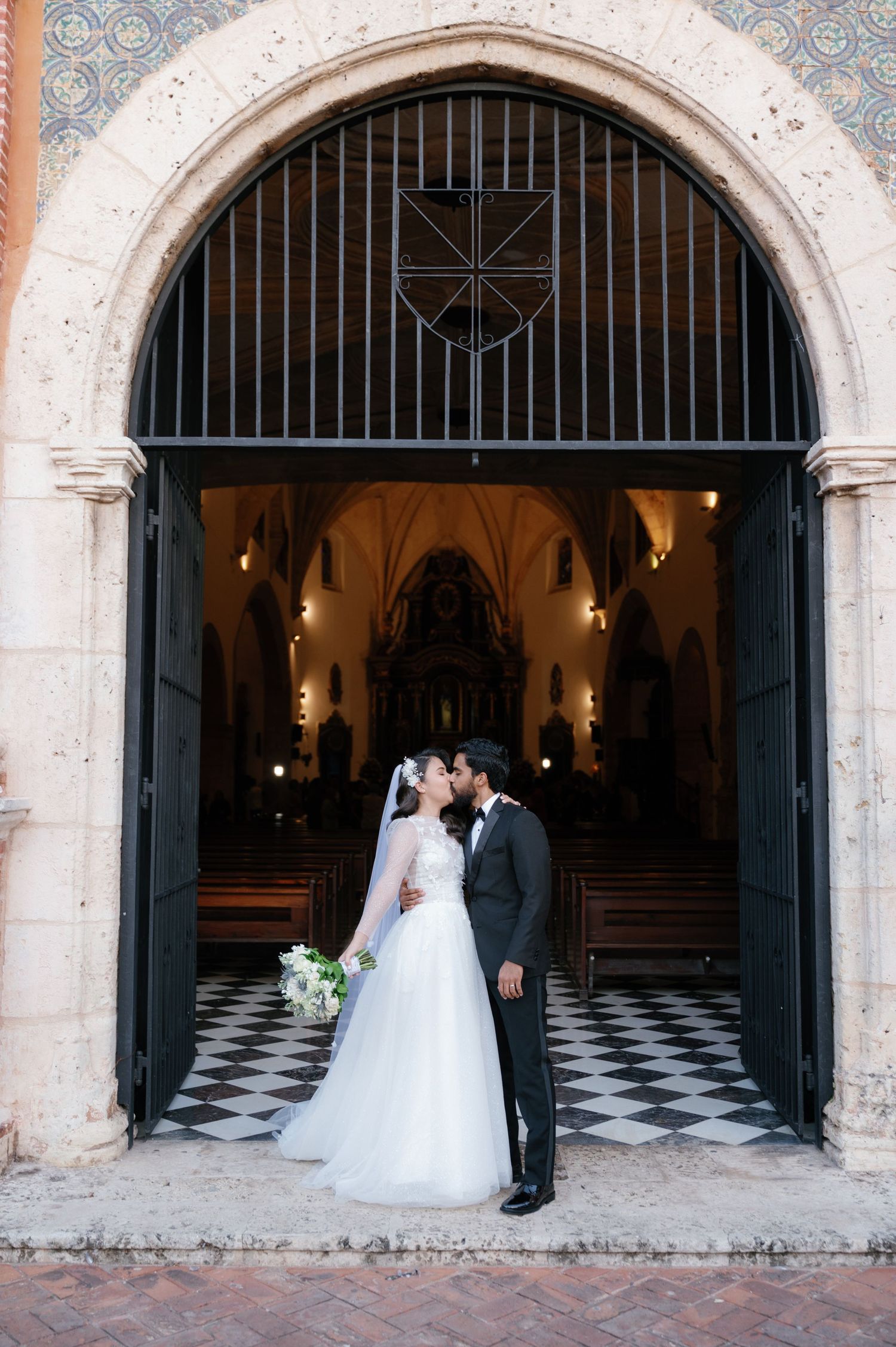 A romantic wedding couple stands beneath stone archways with checkered floors and warm lighting in an elegant venue.