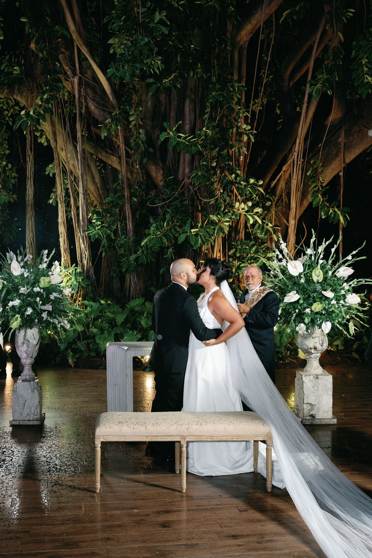 A couple shares a romantic dance moment under banyan trees at a tropical wedding reception decorated with white florals.