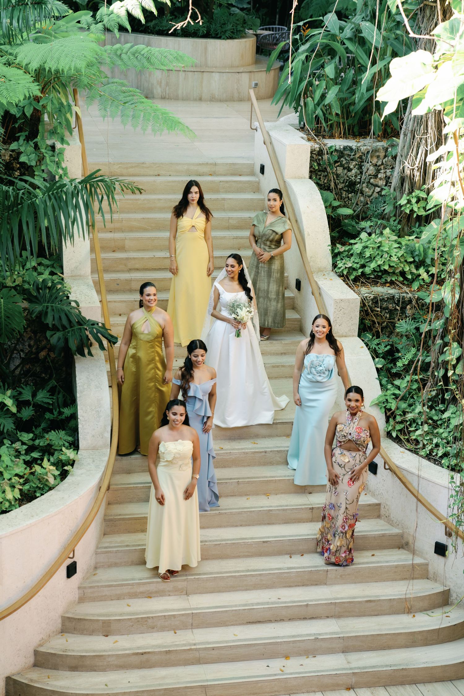 Wedding party members in pastel dresses descending outdoor stone steps surrounded by lush greenery.
